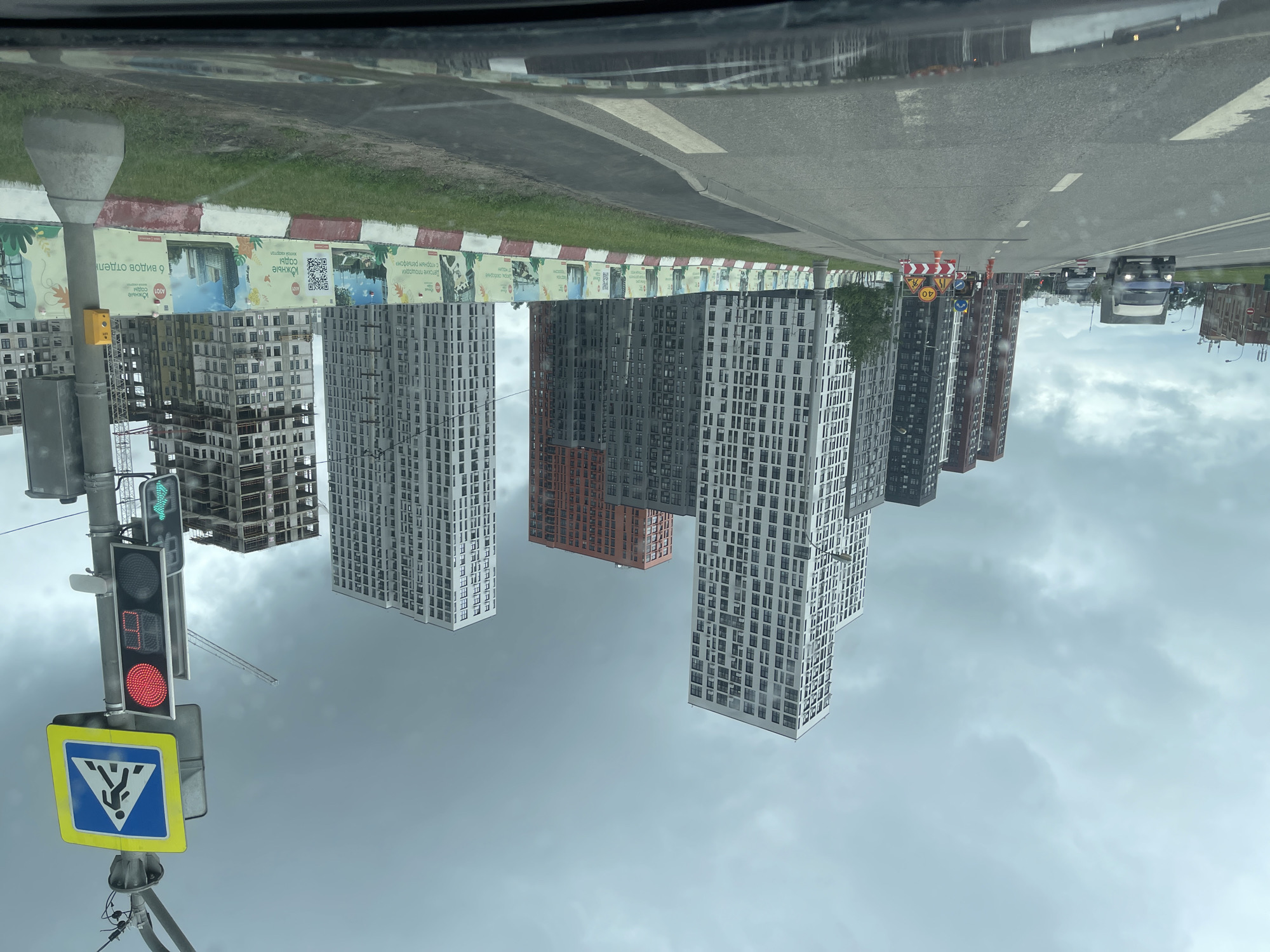 The image depicts a street scene in Moscow, Russia, with several tall, modern residential buildings under construction or recently completed. The buildings are a mix of white, grey, and orange facades, with many windows. The sky is overcast and cloudy.
In the foreground, a road is visible with traffic lanes marked by white lines. A car is driving on the road. To the right of the road, there is a traffic light displaying a red signal and a pedestrian crossing sign. Cones and temporary barriers are set up on the side of the road, indicating roadwork or construction.
A long fence with colorful advertisements or promotional materials runs along the side of the road, separating it from a grassy area and the construction site. The advertisements feature images of buildings, text in Russian, and QR codes.
Several cranes are visible in the background, suggesting ongoing construction activity. The overall impression is a developing urban area with a mix of residential construction and active infrastructure.