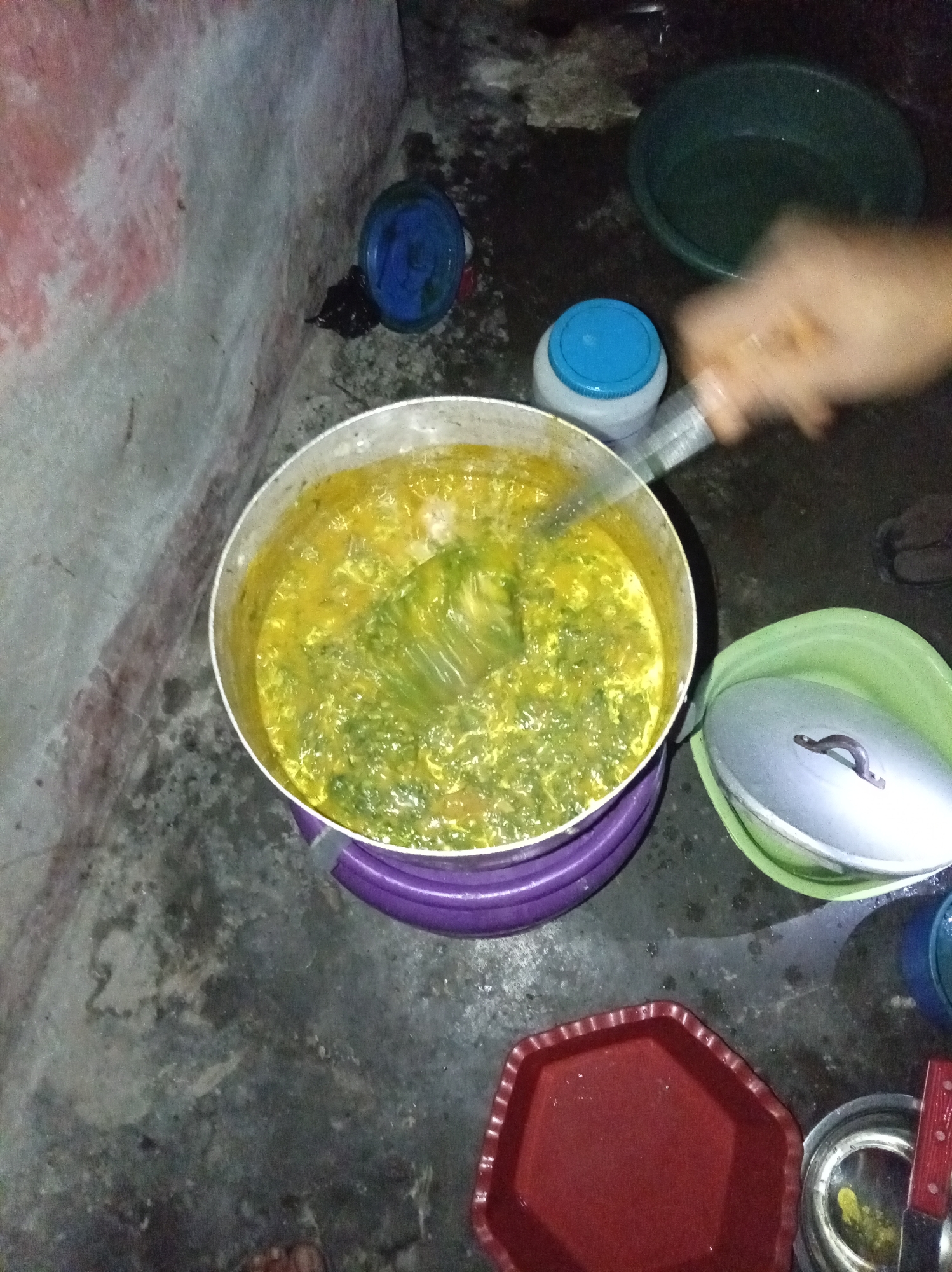 The image shows a pot of soup, possibly being stirred, on the floor of what appears to be a domestic setting in Owerri, Nigeria. The soup is a yellow-green color, containing leafy vegetables. A hand holding a cooking utensil is visible. The pot is placed on a purple ring, possibly a makeshift stand. The surrounding floor appears to be stained or wet, and several plastic containers and other items are scattered around. The background shows a wall with uneven coloring.