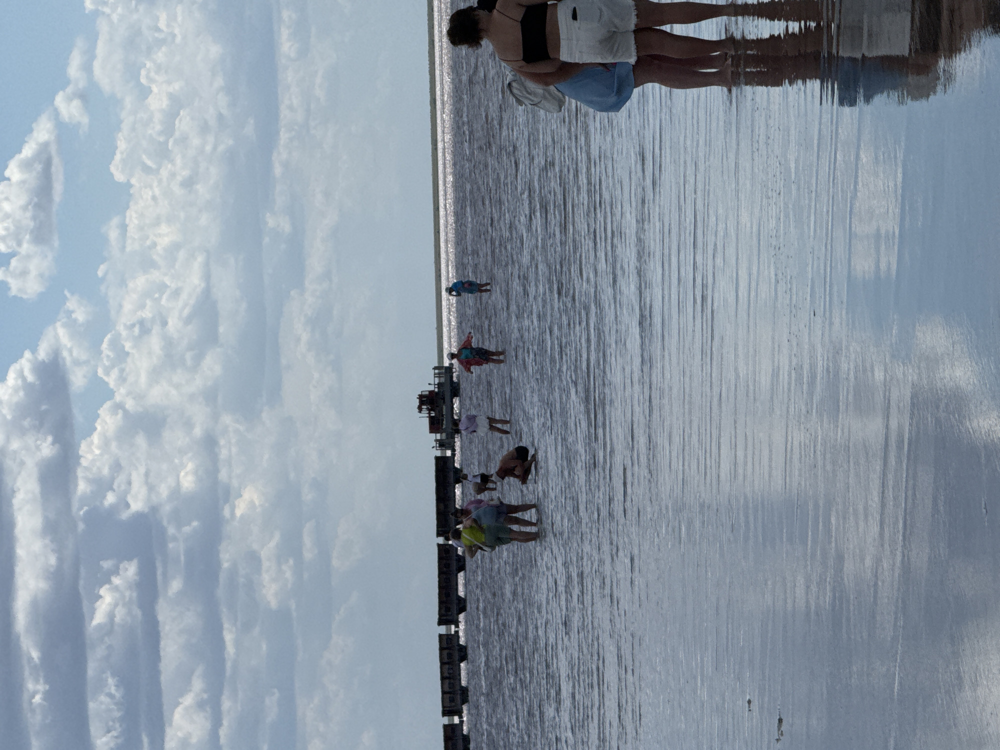 The image shows a group of people wading in a shallow body of water with a pinkish hue, possibly a salt lake, reflecting the sky and clouds. In the background, a train with several carriages and a red tractor on a barge are visible on the water. The sky is filled with dramatic, layered clouds. The scene appears to be taking place in Bursol', Russia, a location known for its salt lakes. The people are casually dressed, some in swimwear and shorts, and they seem to be enjoying the experience of walking in the water. The water is calm with gentle ripples.