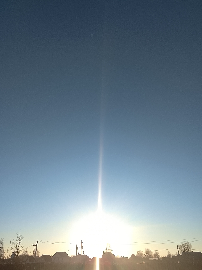 An outdoor, low-angle photograph captured in Высокиничи, Russia, depicts a rural or suburban landscape under a clear, pale blue sky at what appears to be sunrise or sunset. A brilliant, overexposed sun is positioned near the horizon, slightly right of the frame's vertical center, casting an intense, narrow vertical light pillar upwards into the sky. A small, faint white spot is visible in the upper left quadrant of the sky. The lower third of the image is dominated by a silhouetted skyline comprising multiple residential structures with gabled roofs, sparse, bare deciduous trees, and several utility poles with overhead power lines extending horizontally across the scene. No individuals or specific actions are discernible. The ground in the immediate foreground is dark and indistinct due to backlighting.