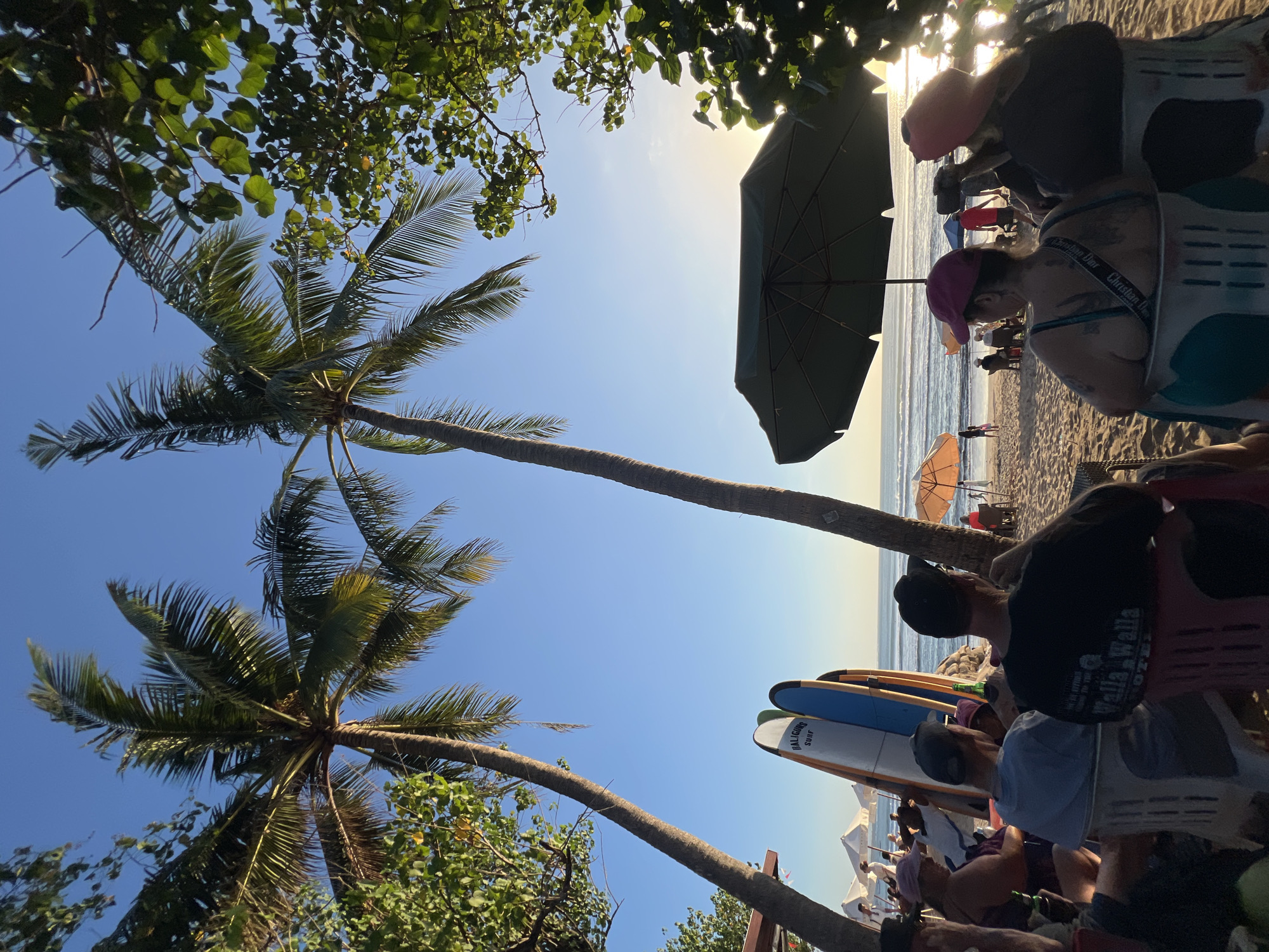 In Kuta, Indonesia, on a beach under a clear blue sky, a group of people are seated on chairs, facing the ocean. Two prominent palm trees frame the scene, with lush green foliage on the right. Several surfboards are propped up near the shore, suggesting a surf spot. In the distance, the gentle waves of the ocean meet the sandy beach where a few other individuals are present. The sun is low in the sky, casting a warm glow and creating a serene atmosphere.