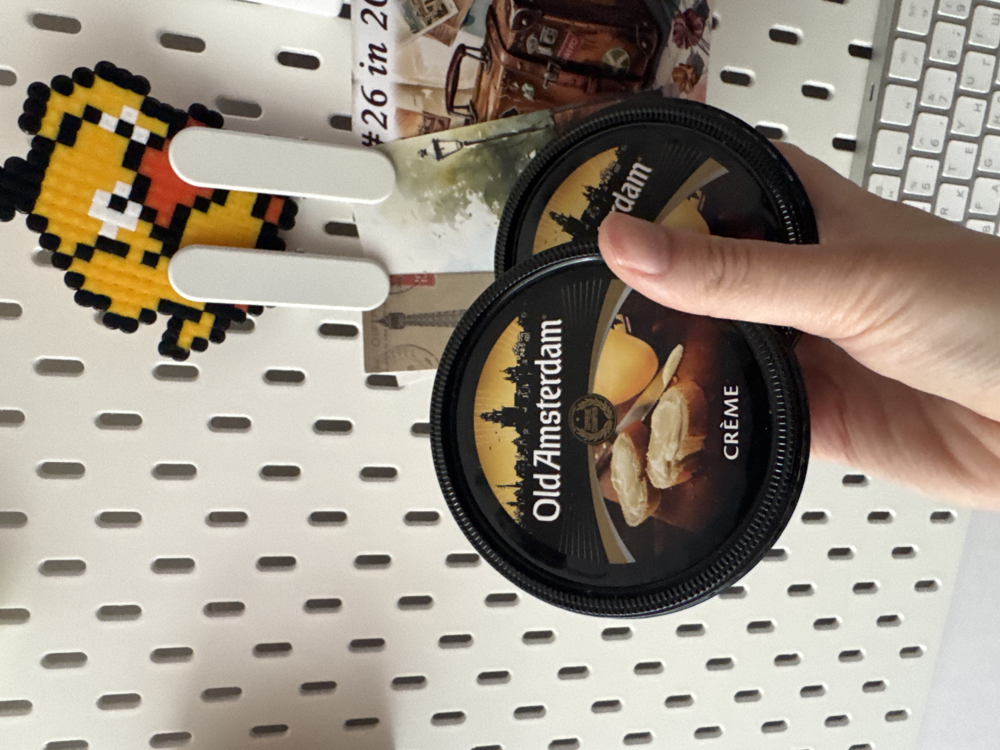 This close-up, angled photograph captures a person's hand holding two food containers against a white pegboard adorned with various items. The setting is an indoor space, likely a desk or personal area, located in Riga, Latvia.
In the foreground, a light-skinned hand holds two round, black plastic tubs of "Old Amsterdam CRÈME" cheese spread. The lids feature a golden silhouette of a city skyline and an appetizing image of cheese and what appears to be bread. The brand name "Old Amsterdam" and the product type "CRÈME" are clearly visible on the tubs.
To the left, a pixelated magnet, resembling a yellow character with black outlines and orange/red facial features, is attached to the pegboard. Two white, rectangular pegs protrude from the board, holding several small decorative cards or postcards. One card prominently displays "#26 in 20" alongside an illustration of a vintage suitcase and a map. Another card features a lamppost, and a faint silhouette of what appears to be the Eiffel Tower is visible on a different card behind the tubs.
The background consists of a white pegboard, characterized by a grid of evenly spaced, horizontal oval-shaped holes. To the right, a portion of a silver or white computer keyboard with QWERTY layout keys (including 'B', '6', 'P', 'O', 'R', 'K', 'E', 'H', 'G', '9') is partially visible, reinforcing the context of a workspace. The overall image presents a collection of personal items and food products in a casual, organized environment.
