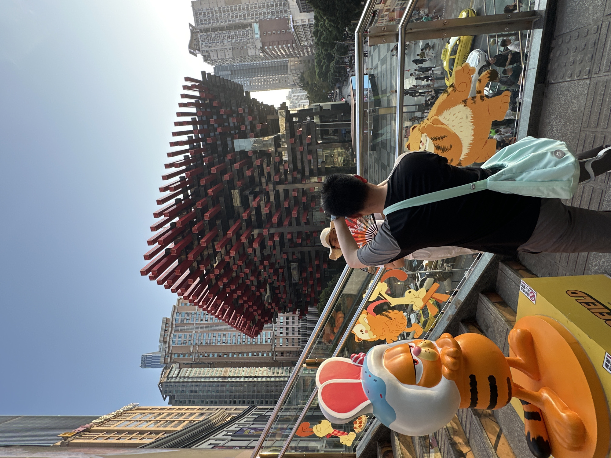A medium, outdoor, canted, eye-level shot depicts a man taking a picture of Raffles City Chongqing in Yuzhong District, China.
The man wears a black shirt, grey pants, and a pale green shoulder bag. He is holding a paper fan in one hand and a phone with the other, taking a picture of the building.
The building is a large, geometric structure made of black and red beams. It is surrounded by tall buildings and a clear blue sky.
In front of the man are several Garfield-themed decorations, including a large Garfield head and a cutout of Garfield lying down. Behind him are other people walking by on the sidewalk.