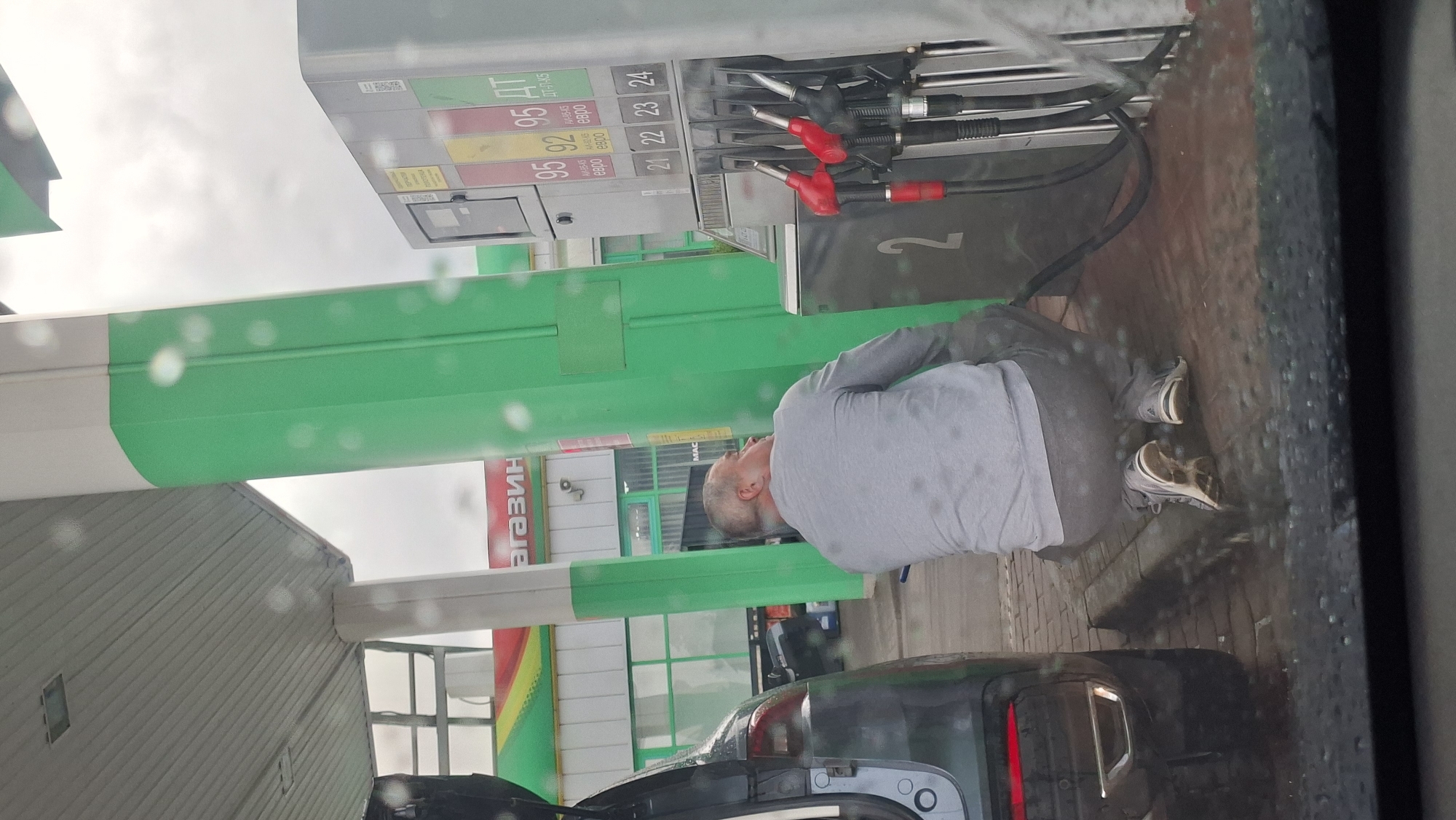 At a gas station in Smalyavichy, Belarus, a man wearing a gray sweatshirt and pants is squatting down next to a car. He appears to be in the process of fueling the vehicle, with a gas hose extending from the pump to the car. The gas pump has various fuel options displayed, including 95 and 92 octane gasoline, and diesel fuel. The scene takes place under a covered area of the gas station, with a green and white pillar visible. The ground is paved with reddish-brown bricks, and there are droplets of water on the surface and on the windshield from which the photo is taken, suggesting it is raining or has recently rained. The background shows a store with the word "МАГАЗИН" (MAGAZIN, meaning "shop" in Russian) visible on its facade.