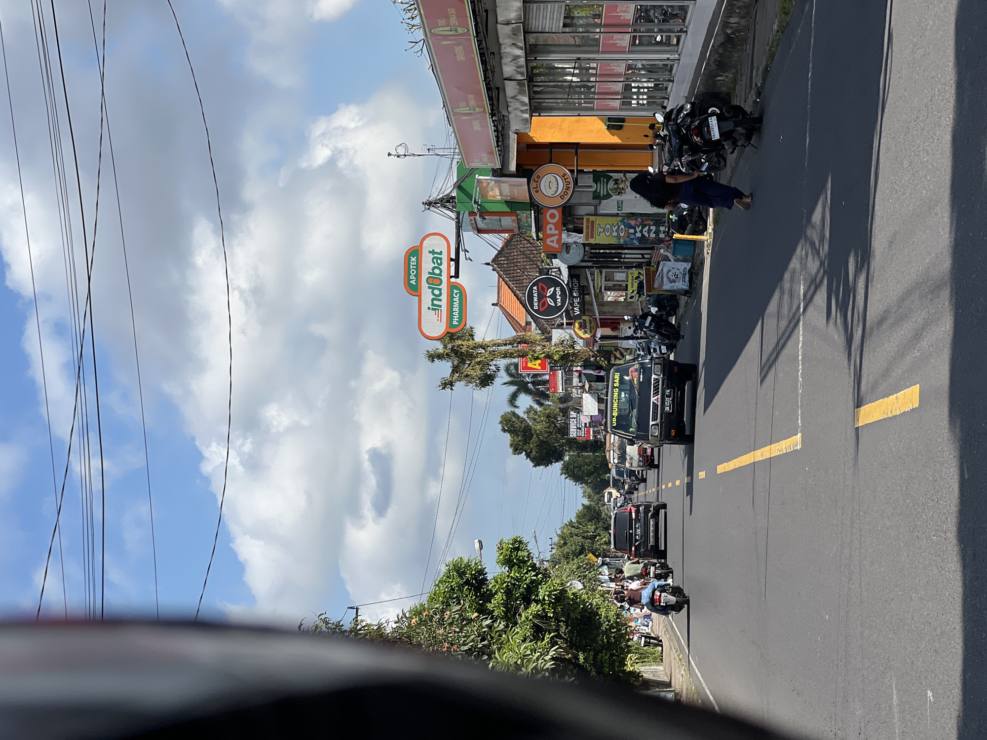 The image depicts a street scene in Tampaksiring, Indonesia, on a sunny day with scattered clouds. The perspective is from a moving vehicle, likely a motorbike, given the presence of a partial helmet visor in the foreground. The street is paved and features a yellow double line marking the center. Traffic is moderate, with several cars and motorbikes on the road.
On the right side of the street, a row of shops and businesses is visible. These include an "APOTEK Indobat PHARMACY" with a distinctive orange and white sign, a vape shop named "DEWATA VAPOR," a donut shop labeled "eL.Co DONUTS," and other businesses with signs in Indonesian. The buildings have a tropical architectural style, with orange and yellow facades and tiled roofs. Many motorbikes are parked along the sidewalk in front of the shops. A woman in a dark blue dress is walking barefoot away from the camera towards the parked motorbikes.
On the left side of the street, lush green trees line the sidewalk. More motorbikes are parked there as well, and a few people are on them. The sky is a bright blue with cumulus clouds. Overhead, a network of power lines is strung across the street. The overall impression is a bustling commercial street in a tropical Indonesian town.