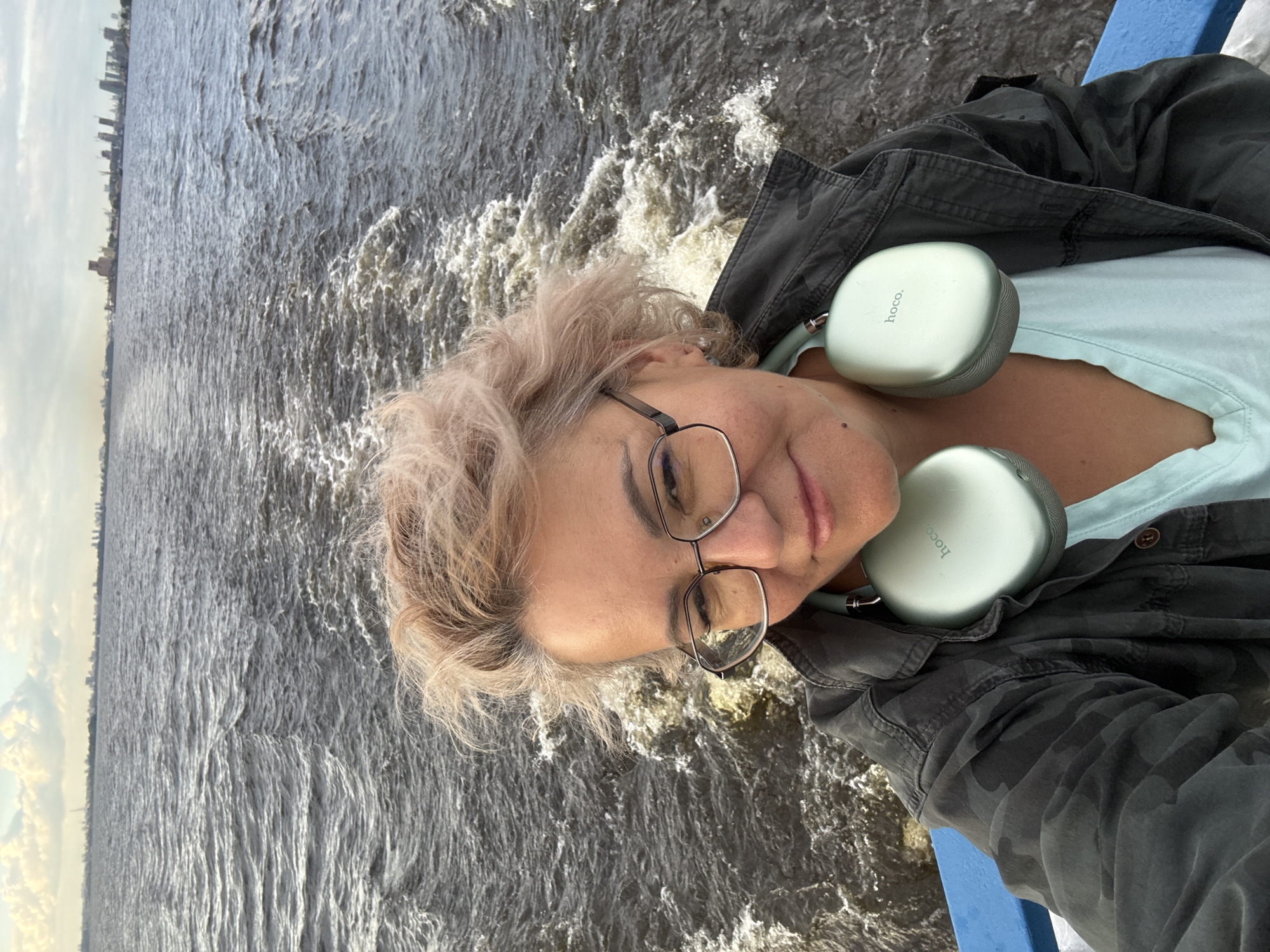 A middle-aged Caucasian woman with blonde, wavy hair and glasses is taking a selfie from a boat on a body of water. She is wearing a dark, camouflage-patterned jacket over a light blue shirt and has light green over-ear headphones around her neck. The water behind her is choppy, with a wake trailing from the boat. In the distance, a city skyline is visible on the horizon under a cloudy sky. This skyline is consistent with the city of Kazan, Russia, featuring industrial structures and buildings. The boat appears to be moving forward, indicated by the wake. The lighting suggests it is daytime, possibly late afternoon given the cloud cover and soft light.
