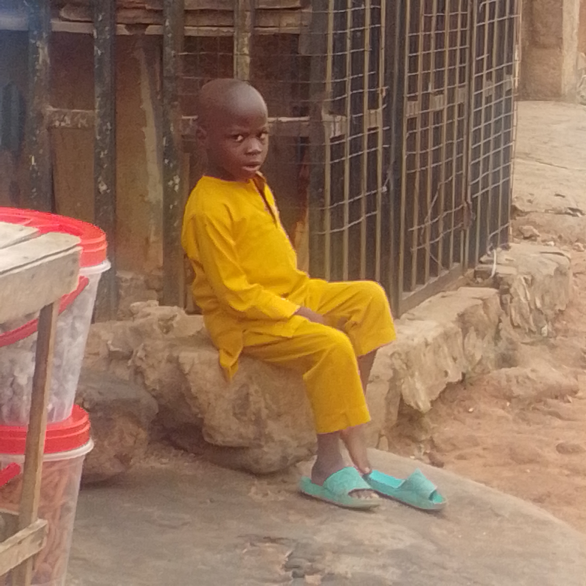 In Tempure, Nigeria, a young boy, appearing to be between 5 and 7 years old, is seated on a rough, textured stone surface. He is wearing a bright yellow long-sleeved shirt and matching loose-fitting pants. On his feet are light blue slide sandals. The boy is positioned in front of a dark metal cage-like structure, possibly a fence or a stall enclosure, made of vertical bars and horizontal crossbars. To the left of the boy, a portion of a stall is visible, featuring two clear plastic containers with red lids, one stacked atop the other. The lower container appears to hold orange-colored items, while the upper one contains lighter-colored objects. The ground is a mix of dirt and small stones. The overall scene suggests an outdoor market or street setting.