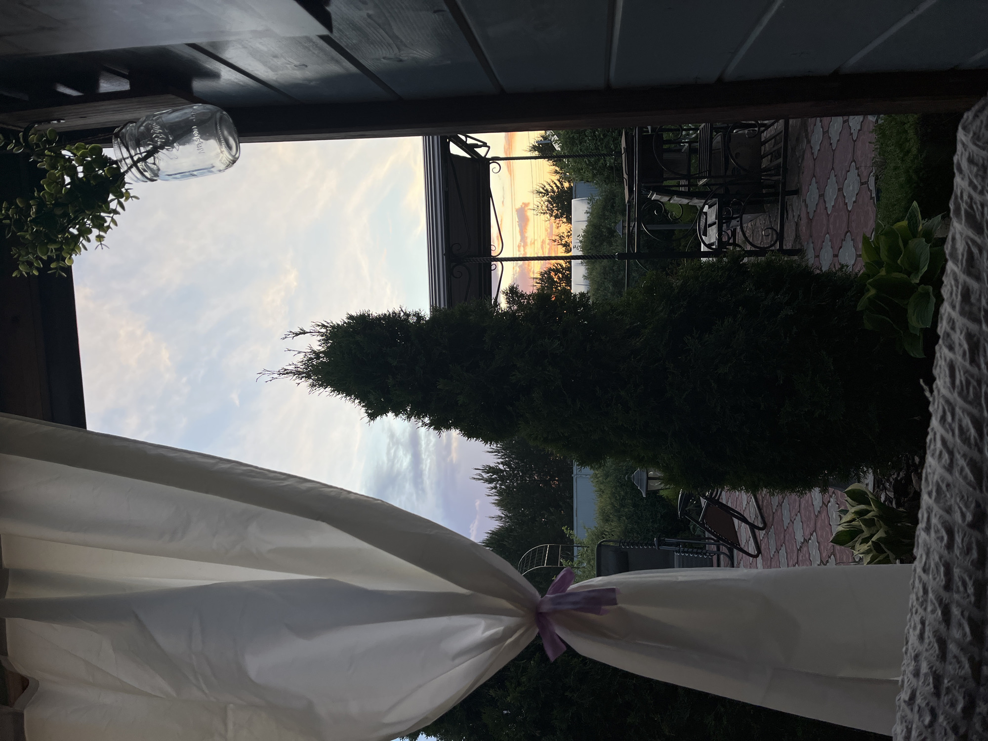 From a sheltered location, the image shows a garden setting at dusk. A tall, conical evergreen is centrally positioned in the view. Garden furniture, including chairs and tables, is visible on a patterned patio. A white curtain, gathered with a light purple ribbon, frames the foreground. A clear glass jar with hanging greenery is suspended from the ceiling of the shelter. The sky is a mix of light blue, purple, and orange hues, indicating either sunrise or sunset. The location is Chaikovsky, Russia.