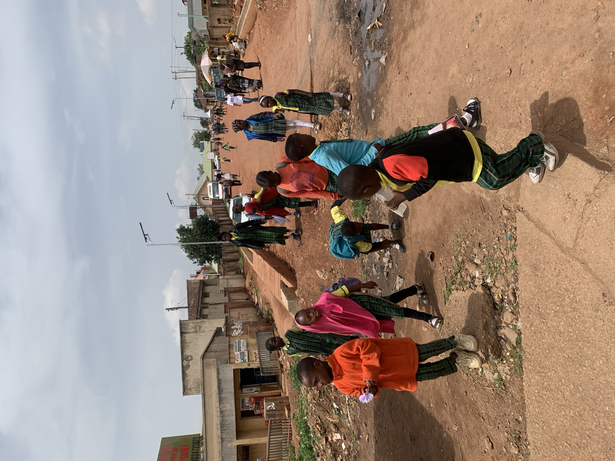 The image depicts a street scene in Gura Top, Nigeria, under a cloudy sky. Several children, some in school uniforms consisting of plaid skirts and green tops, are walking along a dirt road. One child in an orange jacket is holding a small object. Another child in a blue shirt is looking forward. In the background, buildings line the street, and a few vehicles, including a white van, are present. The road is unpaved and shows signs of wear and tear, with some debris scattered along the sides. There are also streetlights visible. The overall impression is a snapshot of daily life in a Nigerian town.