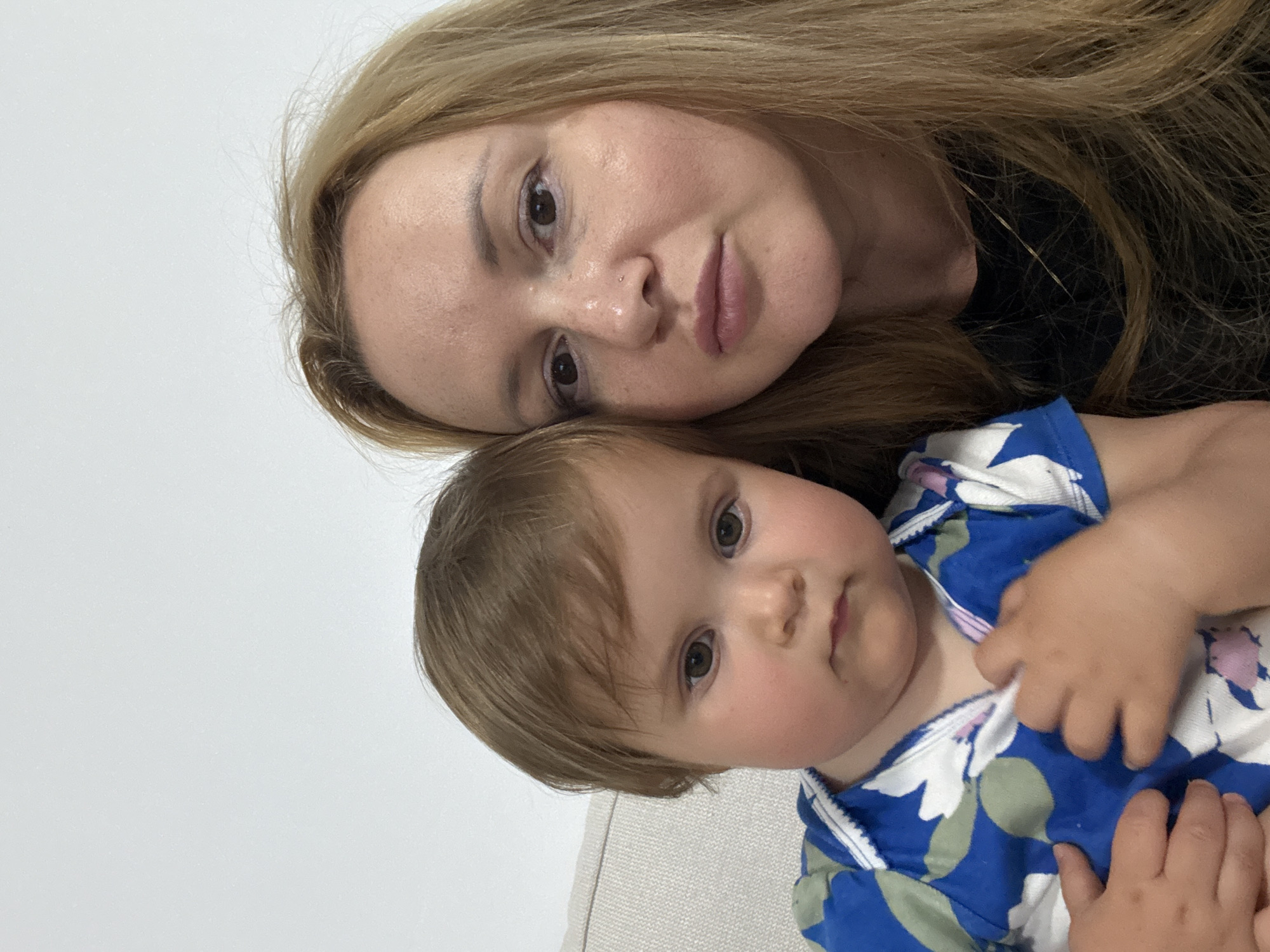 A close-up, rotated portrait shows a woman and a baby positioned intimately together. The image is oriented 90 degrees counter-clockwise, with the subjects appearing to lie down.
The woman, likely the mother, has long, light brown or blonde hair that frames her face. She has fair skin with visible freckles, and brown eyes that gaze directly at the viewer with a gentle expression. Her lips are slightly parted. She is wearing a dark-colored top, barely visible around her hair.
Resting closely beside her is a baby or toddler with light brown hair and wide, dark eyes. The baby has full, round cheeks and a neutral, curious expression, also looking towards the camera. The baby is dressed in a blue top adorned with a white and green floral pattern. One of the baby's hands rests near its chest.
The background is a plain, bright white wall, suggesting an indoor setting. A light-colored, possibly beige or off-white, upholstered surface (like a sofa or bed) is partially visible in the bottom portion of the frame. The lighting is bright and even, casting soft shadows. There is no visible text in the image. The overall mood conveys warmth and familial closeness.
