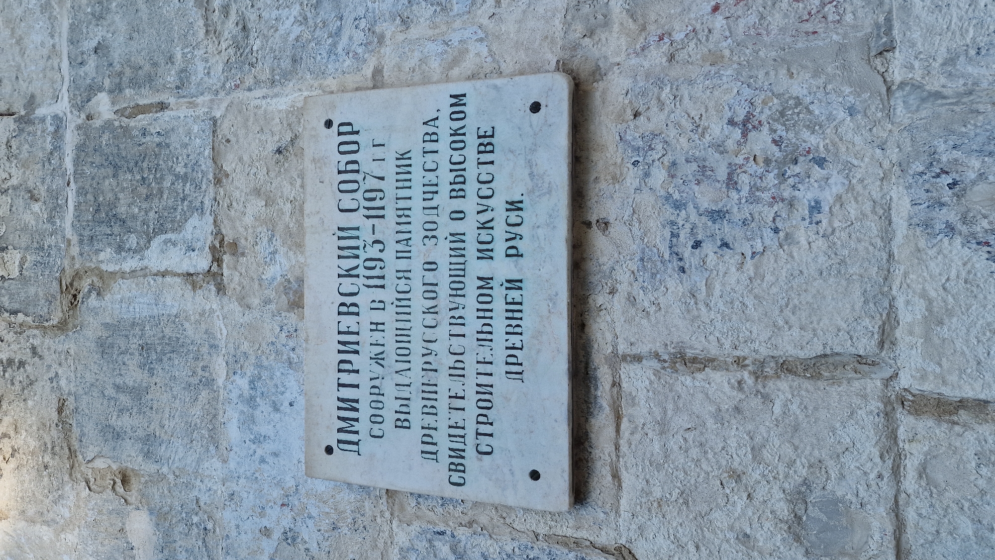This is a vertical, eye-level, medium shot of a historical plaque mounted on a rough, light-colored stone wall. The plaque is off-white and rectangular with rounded corners, and it is oriented vertically, with the text running from bottom to top. The stone wall is textured, with visible cracks, uneven surfaces, and patches of what appears to be ancient plaster or paint in muted shades of blue and red. The lighting is bright, suggesting it is daytime.
The plaque bears an inscription in Russian. The text reads: "ДМИТРИЕВСКИЙ СОБОР СООРУЖЕН В 1193-1197 гг ВЫДАЮЩИЙСЯ ПАМЯТНИК ДРЕВНЕРУССКОГО ЗОДЧЕСТВА, СВИДЕТЕЛЬСТВУЮЩИЙ О ВЫСОКОМ СТРОИТЕЛЬНОМ ИСКУССТВЕ ДРЕВНЕЙ РУСИ." This translates to: "Dmitrievsky Cathedral Built in 1193-1197. An Outstanding Monument of Old Russian Architecture, Testifying to the High Building Art of Ancient Rus'." The plaque is attached to the wall with at least two dark, circular rivets or fasteners. The overall impression is one of historical significance and weathered resilience. The setting appears to be outdoors, likely an exterior wall of an ancient building.