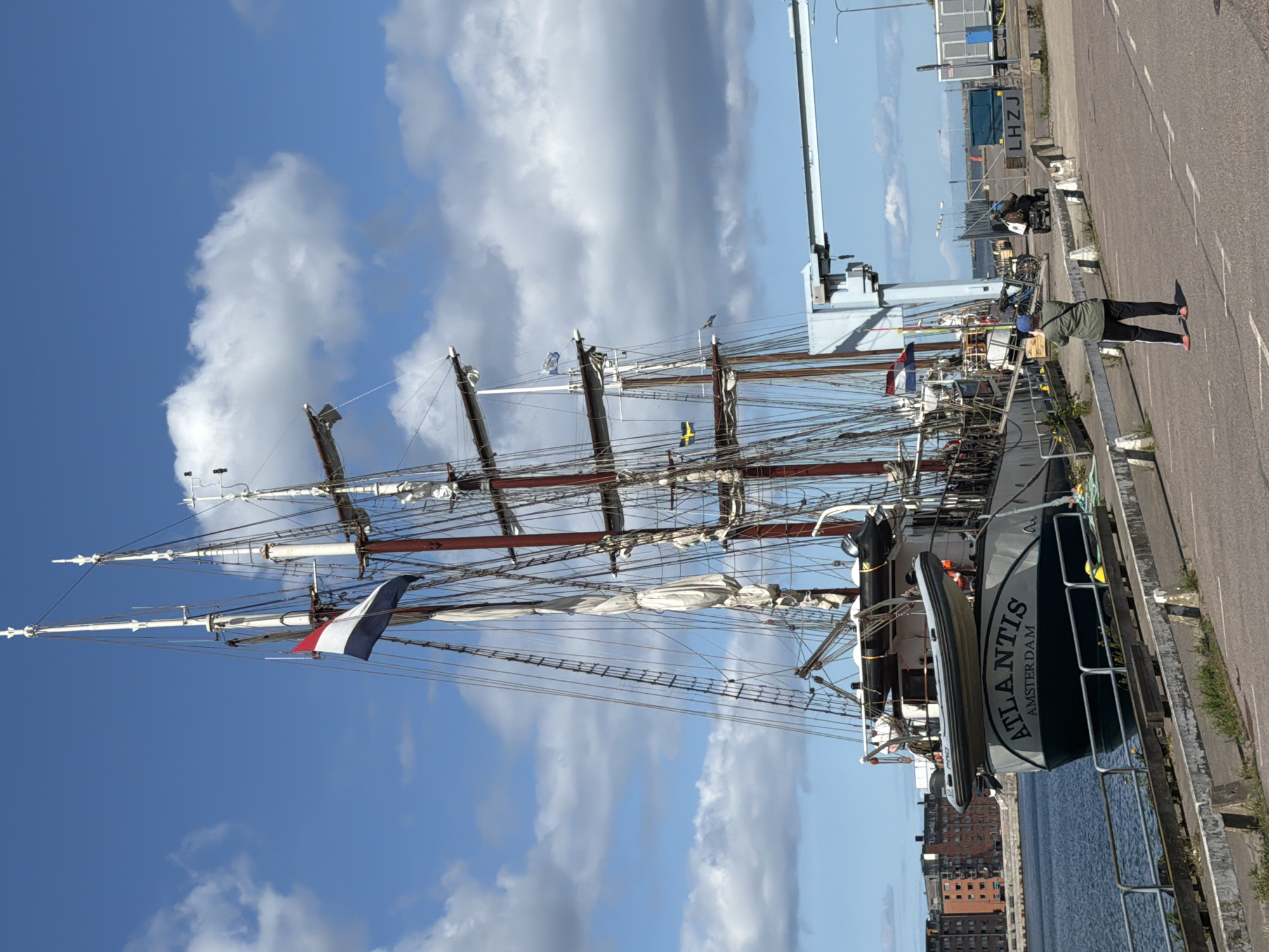 In Malmö, Sweden, a tall ship named "Atlantis Amsterdam" is docked at a harbor. The ship has multiple masts with sails furled and is adorned with a Dutch flag. A person wearing a green jacket and black pants is standing on the dock, facing the ship and holding a camera, seemingly taking a photograph. In the background, industrial structures and buildings are visible. The scene is set under a blue sky with scattered clouds.