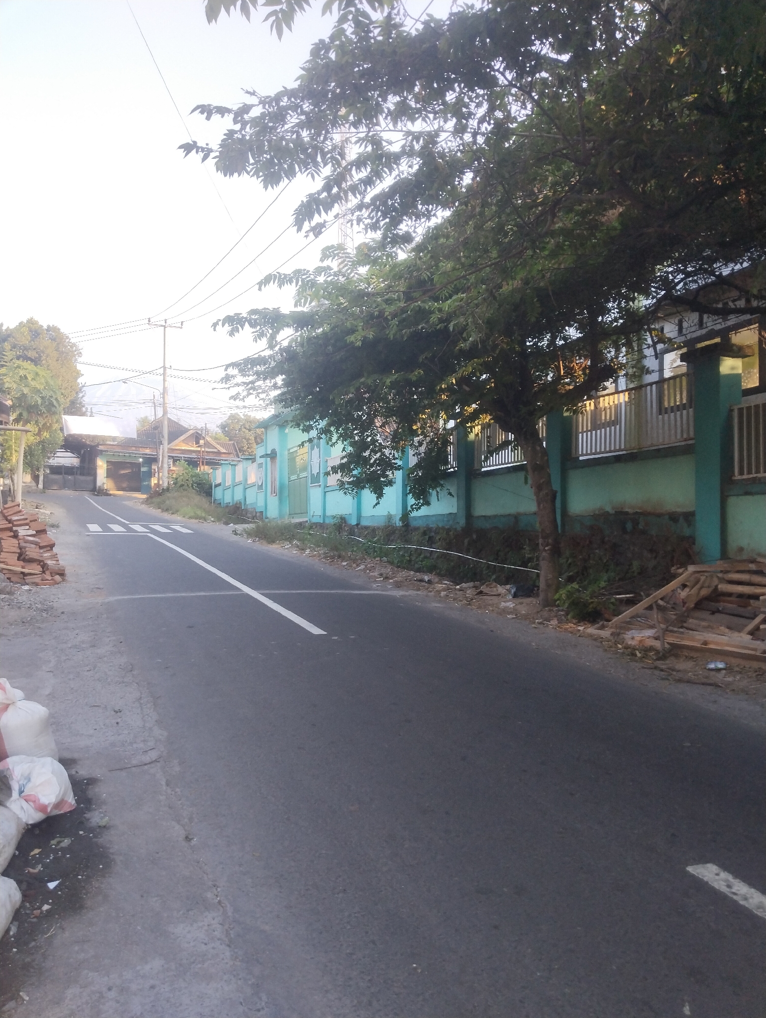 The image depicts a street scene in Wanasaba Daya, Indonesia, with a paved road running through the center. To the left of the road, there is a pile of bricks and several white sacks, suggesting construction or waste materials. The left side of the road also features a sidewalk and some vegetation. On the right side of the road, there is a light blue wall topped with a metal fence. A tree with dark green foliage stands in front of the wall, casting shadows on the pavement. Further down the road, buildings with windows and doors are visible, as well as utility poles with overhead wires. In the distance, mountains can be seen under a bright sky. The overall impression is a residential or semi-rural street with some elements of ongoing work or activity.