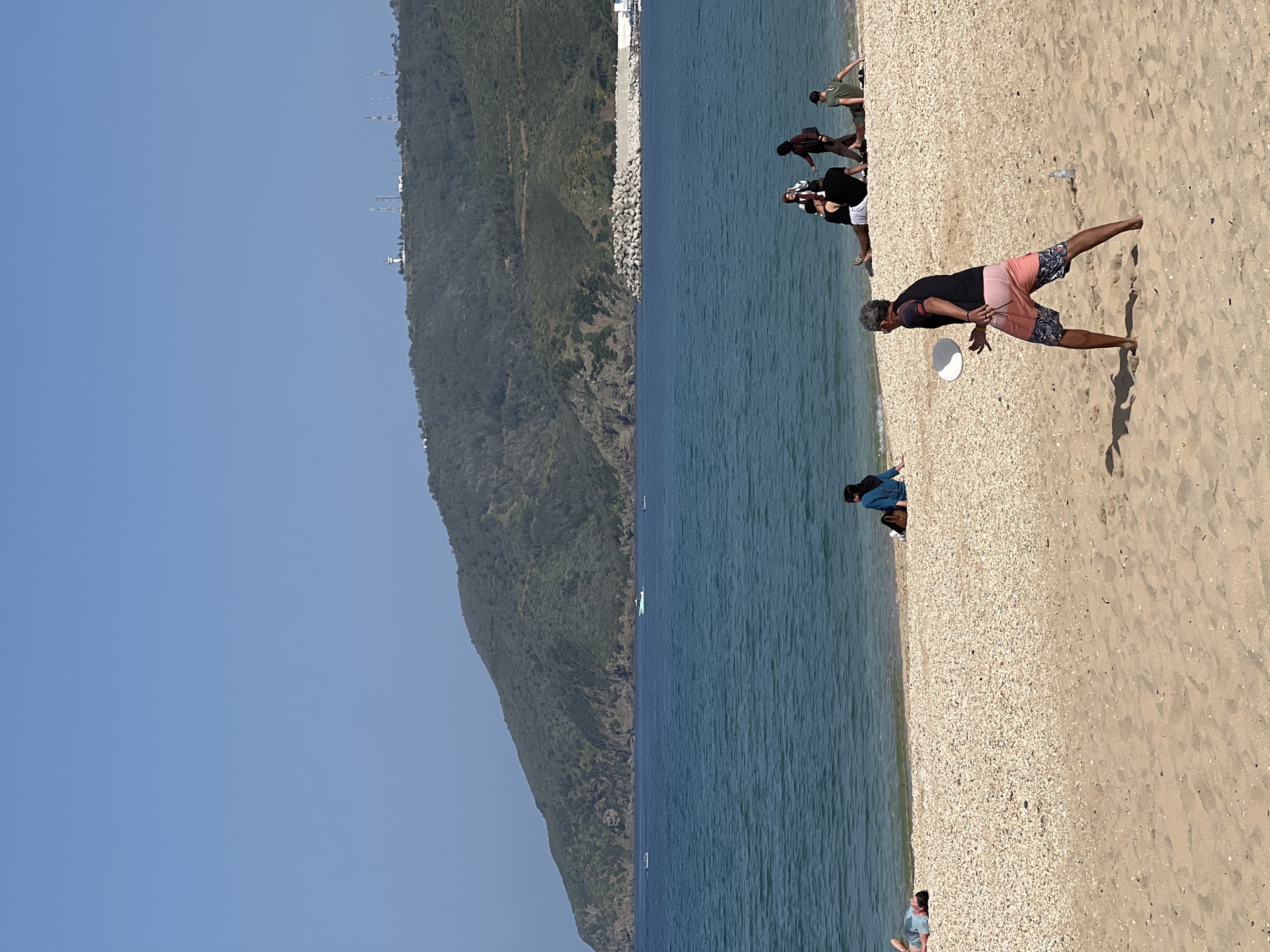 This vertical landscape photograph, taken on a clear and sunny day in M'Diq, Morocco, depicts a lively beach scene. The image is horizontally divided into three main sections: a verdant mountain range on the left, the tranquil blue sea in the center, and a wide, populated beach on the right.
The mountain range on the left is covered in dense green vegetation, extending from the foreground into the distance. Several communication towers or similar structures are faintly visible atop the distant peaks. Along the base of the mountain, concrete tetrapods are arranged, likely serving as a breakwater or coastal defense.
The sea exhibits a clear, deep blue hue with subtle ripples on its surface. A few small boats or buoys are visible in the water in the mid-distance.
The beach on the right is a mix of light-colored sand and small pebbles or shells, particularly near the waterline. Numerous people are enjoying the coastal environment. In the immediate foreground, a man with grey hair and a beard, wearing a black t-shirt and distinctive pink and dark patterned swim shorts, is captured in the act of throwing a white frisbee. His shadow is cast prominently on the sand. Further along the beach, other individuals and small groups are visible: one person in a green shirt, another group of three near the water, and a person in a blue jacket. A discarded plastic water bottle lies on the sand in the mid-ground.
The sky above is a bright, cloudless blue, suggesting good weather conditions. No discernible text is visible in the image.