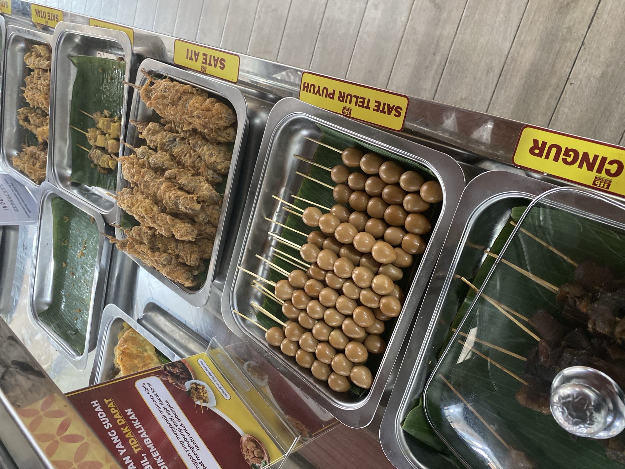 The image shows a close-up, high-angle shot of a food display counter, likely in a restaurant or food stall. In the foreground, a metal tray contains numerous small, hard-boiled quail eggs skewered on wooden sticks, arranged in rows on a bed of green banana leaves. To the left and slightly behind this tray, another metal tray holds pieces of dark meat on skewers, also on banana leaves, with a portion of a glass lid visible.
Above and to the right of the quail eggs, a larger metal tray is filled with fried skewers of food, appearing to be some kind of meat or seafood coated in a crispy batter. Further back, another tray displays skewers with what appear to be cubes of food, possibly meat or tofu, also on banana leaves. The food is presented in rectangular stainless steel trays, common in buffet-style or ready-to-serve food establishments.
Yellow labels with red text are affixed to the front of the display unit. One visible label reads "SATE TELUR PUYUH," which translates to "Quail Egg Satay." Another label partially visible above it reads "SATE ATI," meaning "Liver Satay." A third label at the bottom right reads "CINGUR," a Javanese delicacy made from cow's snout. A red and yellow sign with text in Indonesian is also visible on the left, with phrases like "YANG SUDAH BIL, TIDAK DAPAT DIKEMBALIKAN" (What has been taken cannot be returned) and "Jangan yang mengambil makanan lebih, dapat menghungi staff, agar dapat Kami bantu untuk dibungkus" (If you take more food, please contact staff, so we can help you pack it).
The background consists of a light-colored, possibly wooden, floor and the side of the food display unit, which has a reflective metallic surface. The overall impression is of a bustling food service area, possibly in North Jakarta, Indonesia, given the Indonesian language on the signage. There are no people visible in the image.