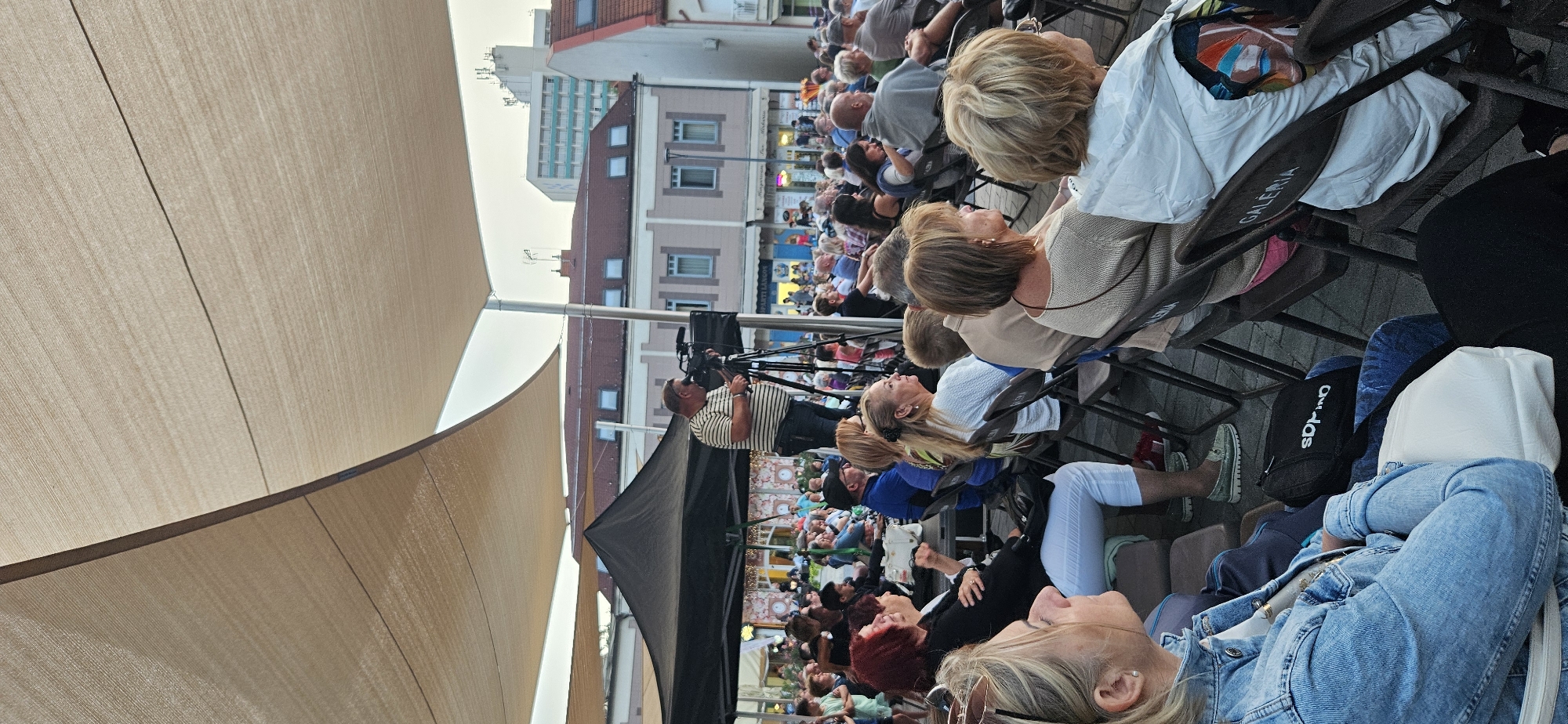 This image captures an outdoor event in Hévíz, Hungary, likely a performance or public gathering. A crowd of people is seated on chairs, facing towards a central area where a man is operating a video camera on a tripod. The man in the striped shirt is focused on his work, suggesting the event is being recorded. The audience is diverse, with individuals of various ages and genders, mostly looking towards the unseen focal point of the event. The setting appears to be a public square or a designated outdoor venue, indicated by the paved ground, the arrangement of chairs, and the buildings visible in the background. A large, tan-colored canopy provides shade over the seating area, and a smaller black tent is also present, possibly for performers or equipment. The architecture of the background buildings, with their pitched roofs and window styles, is characteristic of Central European urban areas. The overall atmosphere seems to be one of attentive observation and engagement with the ongoing event.