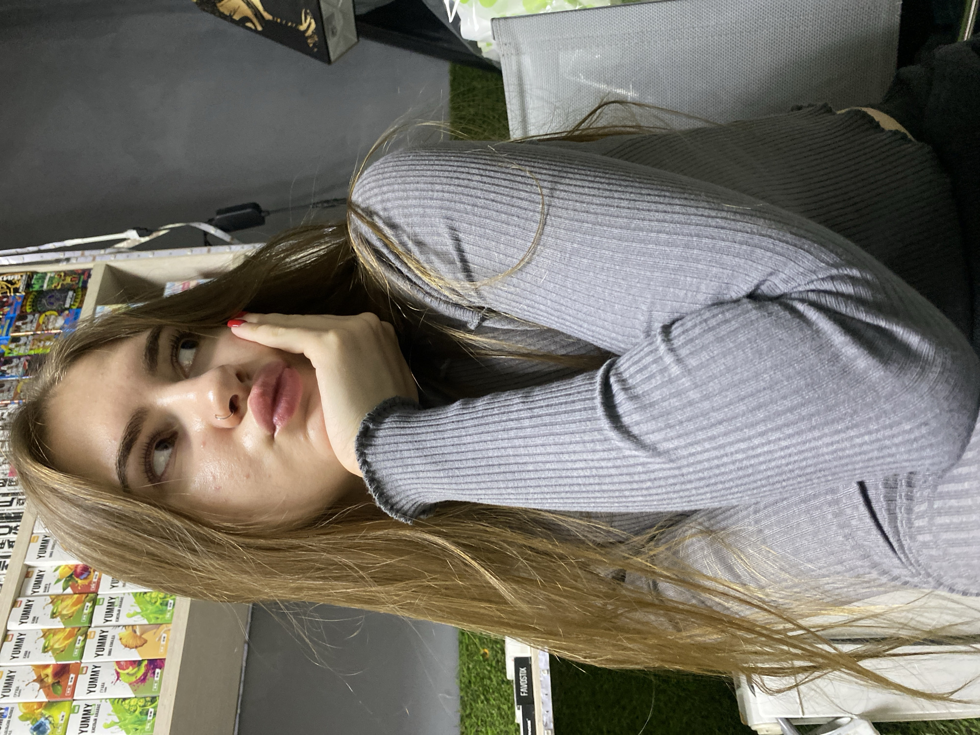 A young woman with long, blonde hair is captured in a medium shot, leaning her head on her hand. She is wearing a grey, ribbed, long-sleeved shirt. Her lips are pursed, and she is looking to her right, with her right cheek resting on her palm. A subtle nose ring is visible. In the background, shelves are lined with colorful boxes, some labeled "YUMMY." To the far right, a dark, textured wall is visible, with a portion of a decorative item with gold accents. A glimpse of artificial green turf is present on the left side of the image, and a portion of a grey chair is visible on the right. The scene appears to be an indoor environment, possibly a shop or a room with merchandise displayed. The location is identified as Dyakovo, Russia.