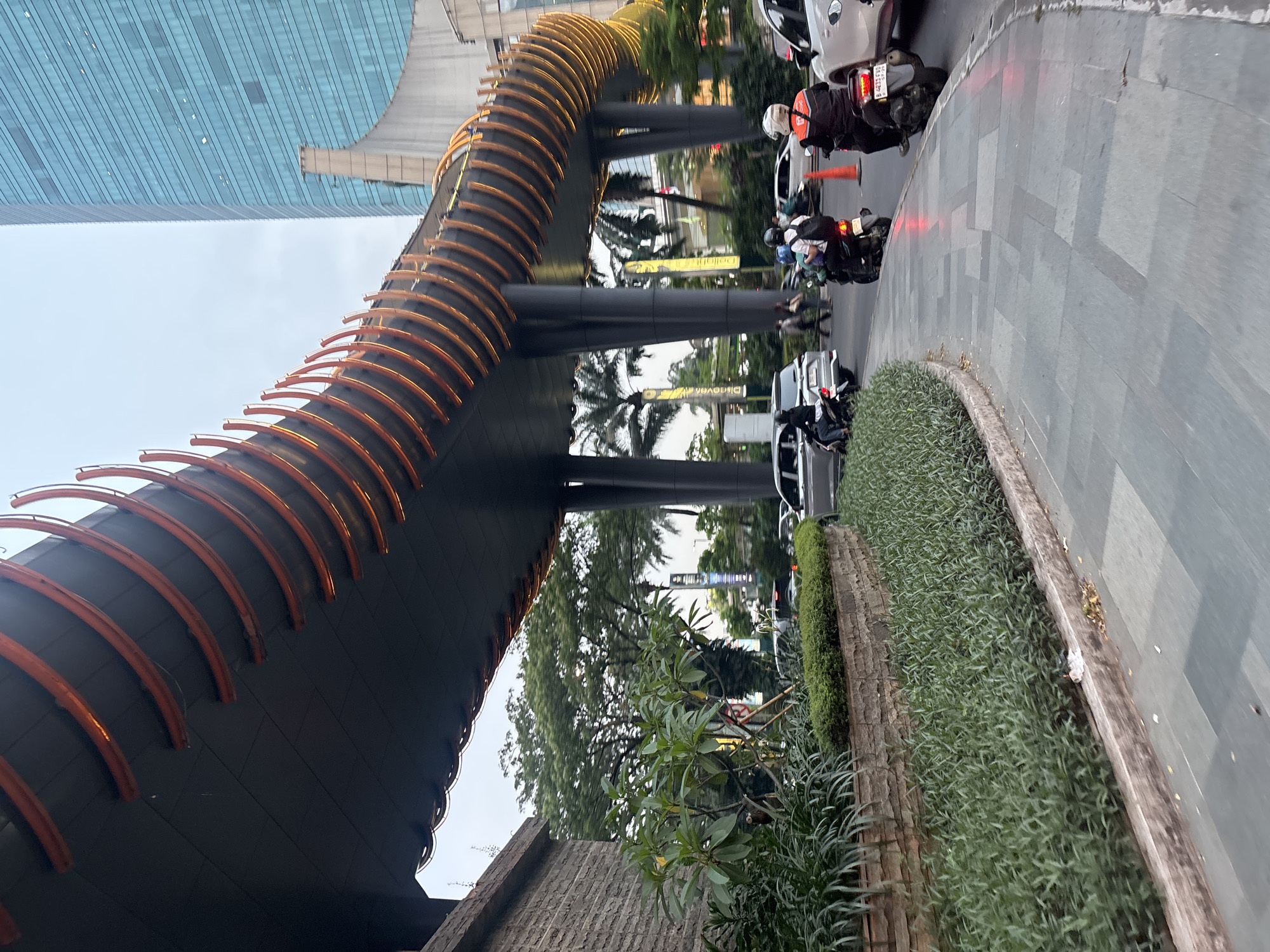 The image depicts an elevated walkway with decorative orange lighting, spanning over a city street in West Jakarta, Indonesia. The walkway is supported by dark pillars. To the left, a building with a stone facade is visible, with lush green foliage in the foreground, including trees and bushes. The street below is busy with traffic, including several motorcycles and cars. Pedestrians are also present, walking on the sidewalk. In the background, a modern, tall building with a blue glass facade dominates the skyline, indicative of a central business district. The lighting suggests it is either early morning or late afternoon, with soft natural light.