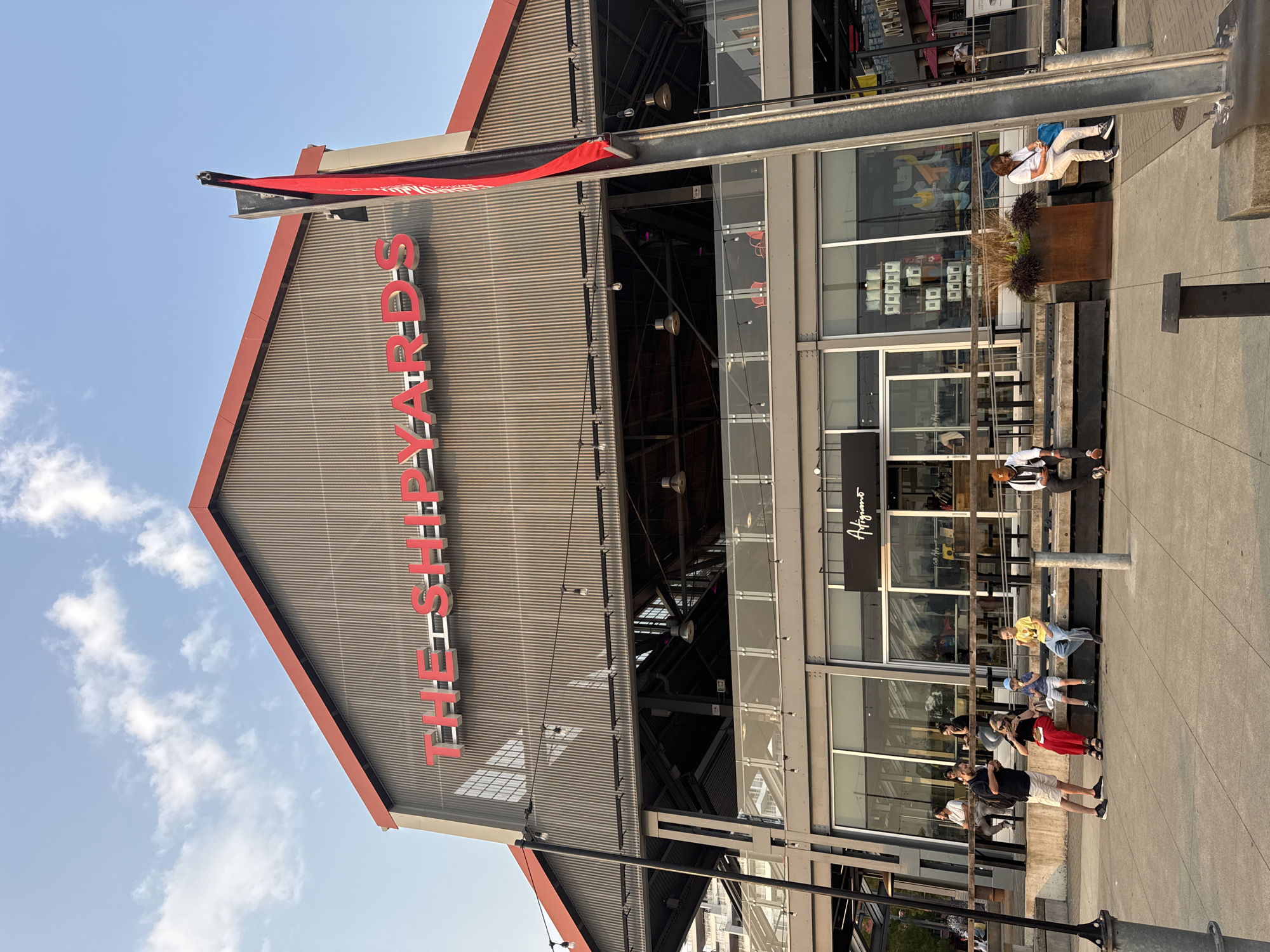 The image shows the exterior of a building with large red letters spelling "THE SHPYARDS" across its facade. The building has a corrugated metal exterior with a red roof. Below the large lettering, there is a covered outdoor seating area with tables and chairs visible through glass railings. The ground level features large windows and doors, with a sign reading "Artigiano" above one entrance. Several people are visible in the foreground and midground, some sitting on wooden benches along the waterfront, while others stand and converse. One person is seated on a bench to the right, looking at a blue bag. The scene appears to be a public space with a clear sky and sunlight casting shadows. This location is The Shipyards in North Vancouver, Canada, a waterfront development known for its public spaces, restaurants, and historical significance.
