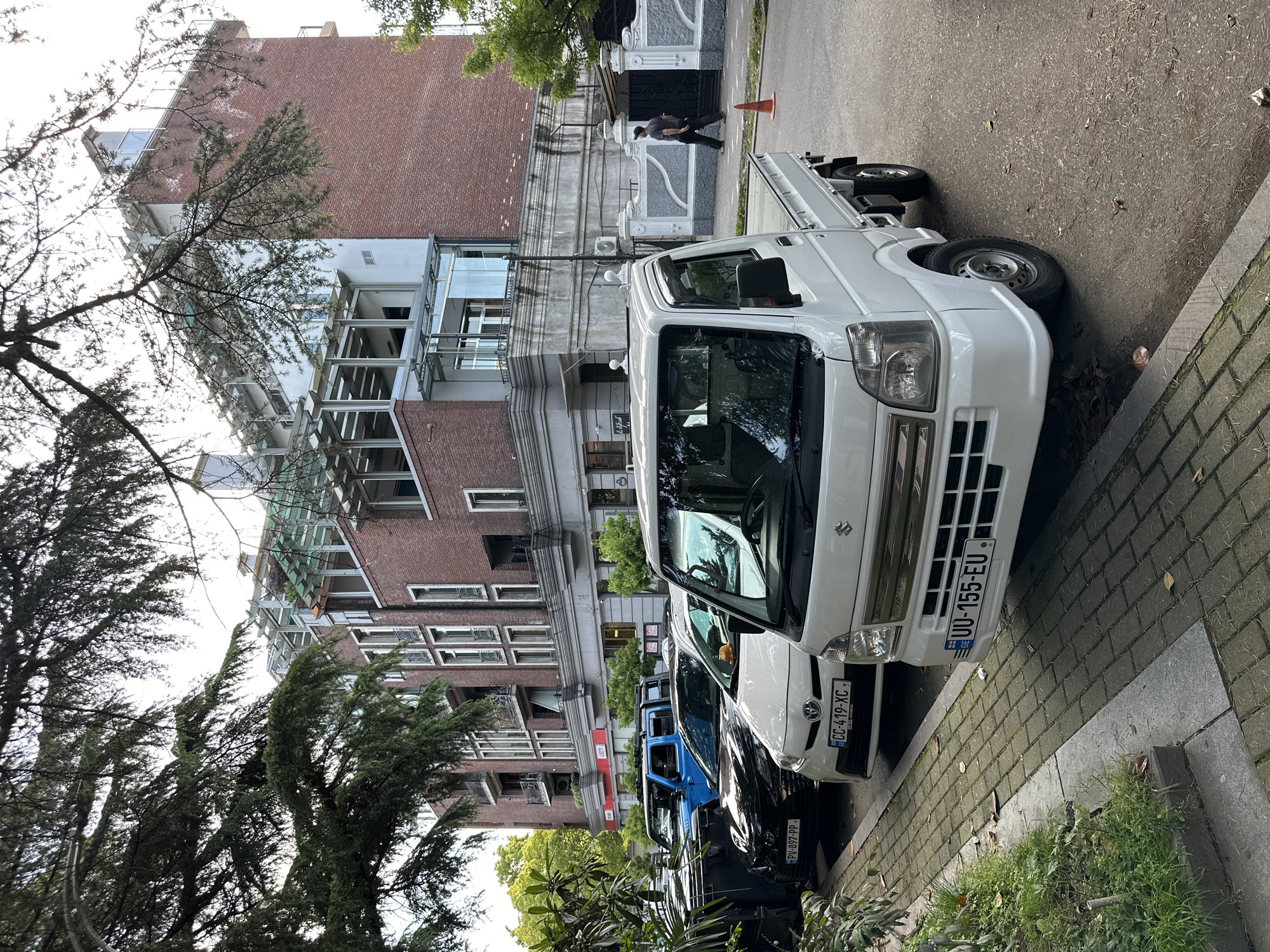 The image depicts a street scene in Batumi, Georgia. The primary subject is a white Suzuki Kei truck parked on the side of the road, displaying the license plate "UU-155-EU." Adjacent to it are other vehicles, including a white Toyota with license plate "CC-419-XC GEORGIA" and a dark-colored car bearing the license plate "PV-897-PP." A multi-story building with a brick facade and modern glass extensions forms the backdrop, partially obscured by trees. A person is walking on the sidewalk. The street has a brick-paved curb.