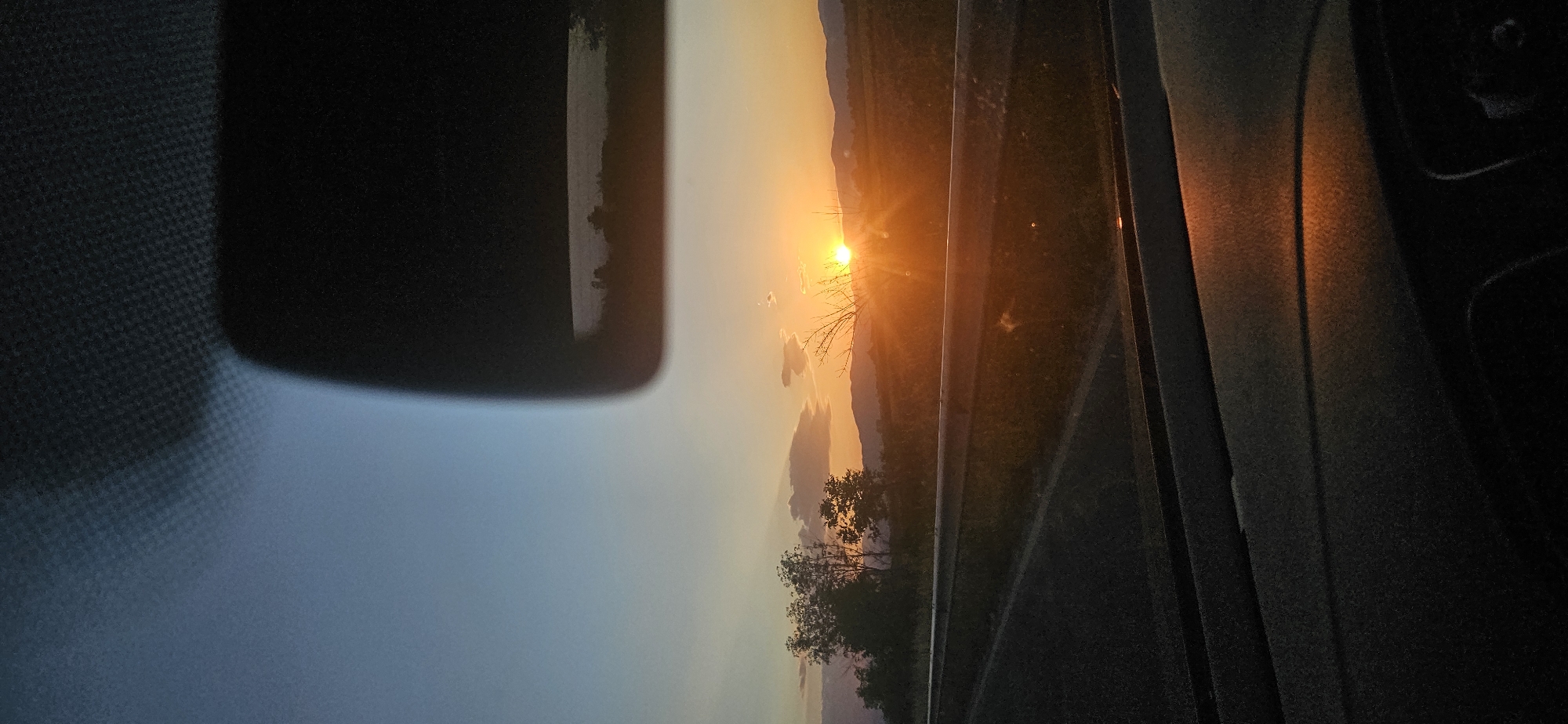 A sunset is viewed from inside a vehicle. The sun is visible over a landscape with trees and a road. A rearview mirror is partially visible in the top left of the frame, and part of the interior of the car is visible on the right. The location is Kovachevo, Bulgaria.