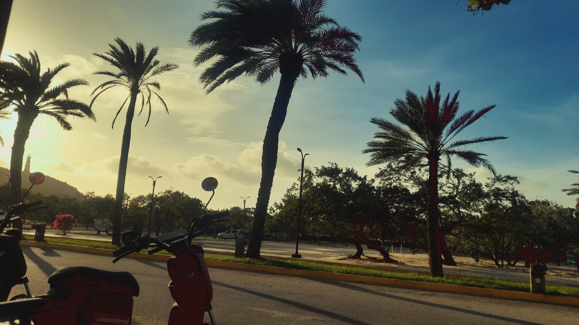 The image depicts a street scene in Pedro González, Venezuela, likely during sunset given the warm, golden light. Several palm trees are silhouetted against a bright sky with scattered clouds. In the foreground, parts of two red and black scooters are visible, suggesting a rental service is present, indicated by the "Happy Rental" text on one of the scooters. The road appears to be asphalt, bordered by a yellow line and a grassy median with a few trees. In the background, a hill with a lighthouse-like structure is visible on the left. The overall atmosphere is tropical and serene, with a hint of urban infrastructure.