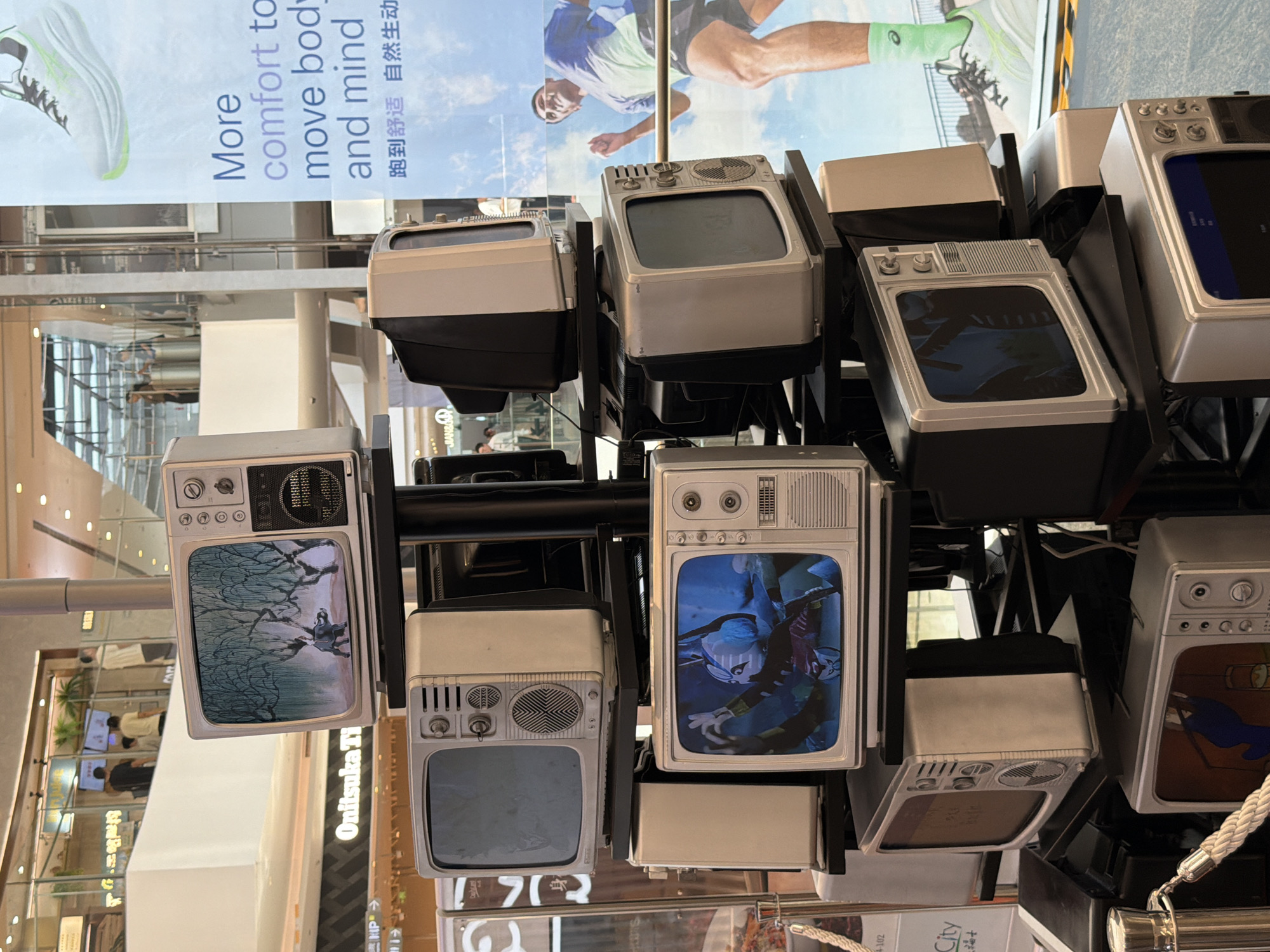 This is a medium-shot, eye-level, indoor photograph of a display of vintage televisions in what appears to be a shopping mall. The televisions are stacked on shelves and some are displaying content. The television on the top left shows a painting of a dark figure with willow trees in the background. The television below it is black and has a blank screen. The television to the right of that one is also black and has a blank screen. The television in the lower left displays a cartoon image of a character. To the right of that, another television shows a black screen. The bottom-most television shows a dark screen. In the background, there is a large advertisement featuring a runner and text in English and Chinese. People are visible in the background on a higher level of the mall. The overall environment suggests a modern commercial space with escalators and retail displays. The location is identified as Yuzhong District, China.