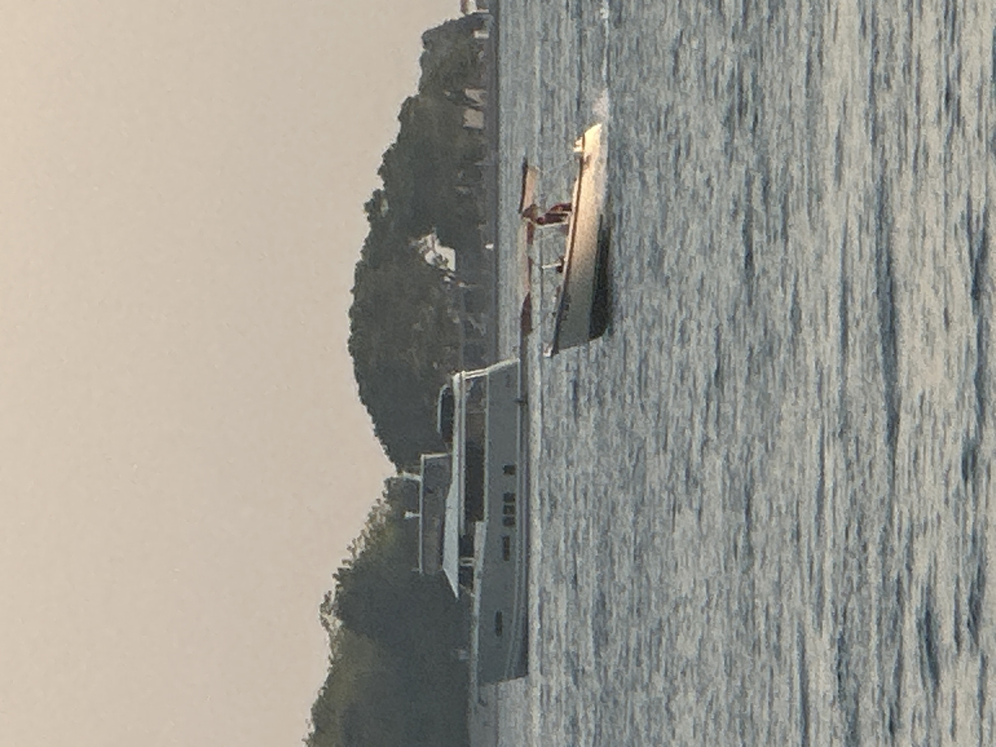 The image captures a serene coastal scene, likely near Budva, Montenegro, with a calm sea reflecting the soft, overcast sky. Two boats are visible on the water. In the foreground, a smaller, lighter-colored open boat with a canopy is moving from right to left, leaving a wake. A person is seated in this boat. In the middle ground, a larger, grey motor yacht is docked or anchored parallel to the shore. Behind the boats, a densely wooded, hilly island or coastline forms the backdrop, with a few indistinct structures visible on its slopes. The water is choppy, with small waves rippling across its surface. The overall atmosphere is peaceful and tranquil.