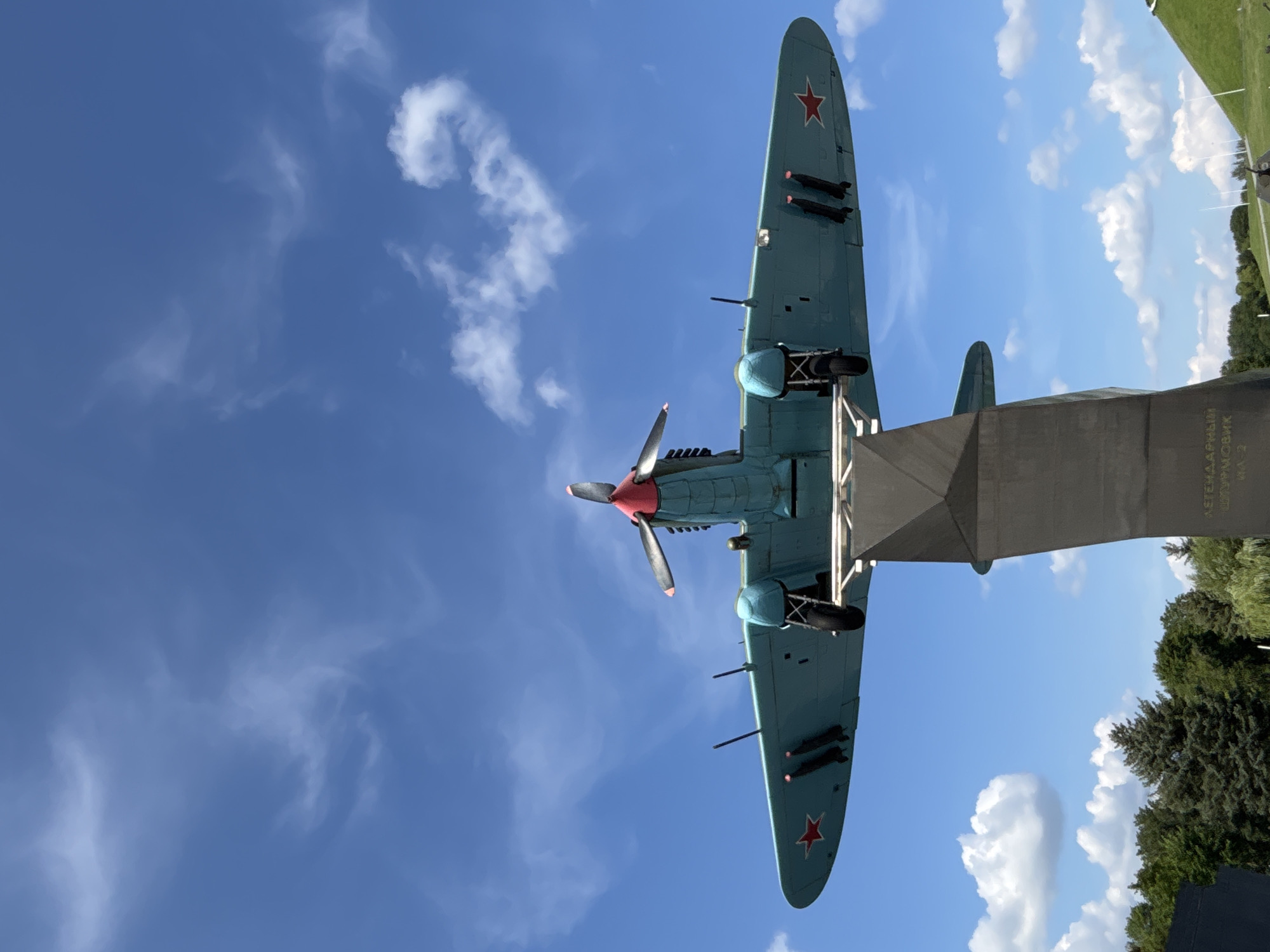 An aerial monument, likely a Soviet-era ground-attack aircraft, is mounted on a concrete pedestal against a bright blue sky with scattered white clouds. The aircraft, painted a light blue with red accents on the propeller spinner and wing insignia, is viewed from directly underneath. It has two wings with visible bomb racks carrying what appear to be four bombs. The cockpit canopy is visible, along with landing gear. The pedestal has some text inscribed on it, possibly indicating it's a memorial. The background includes a line of green trees and a grassy hill on the right side. This monument is located in the city of Ozyaritska-Slabadski rural council, Belarus.