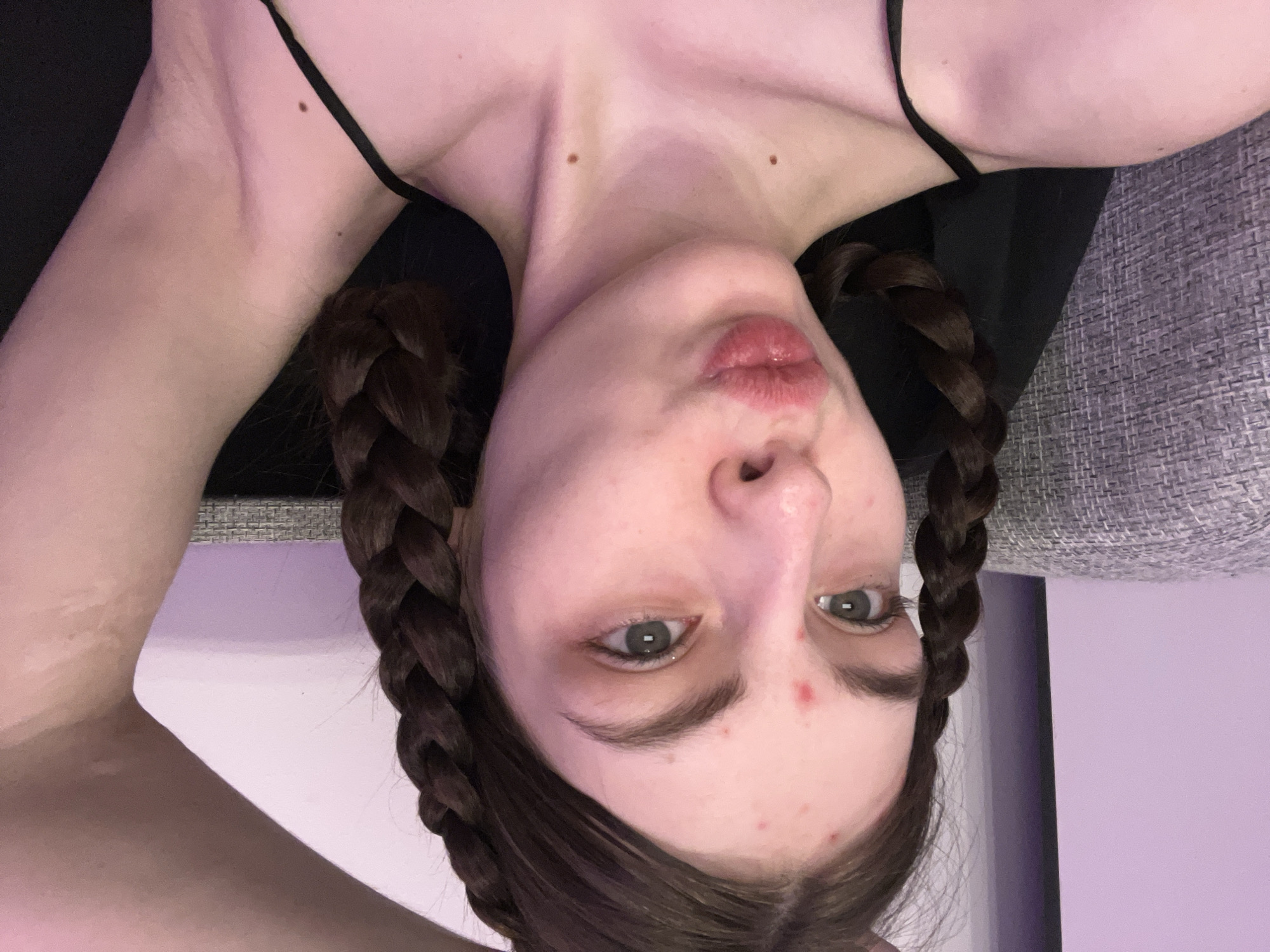 A woman with dark hair braided into pigtails is the central figure of the image. She is wearing a black top with thin straps. Her facial features include slightly pursed lips, green eyes, and some blemishes on her forehead. Two moles are visible on her upper chest. The woman is positioned with her head downwards, giving an upside-down perspective. The background contains a gray textured surface, possibly a couch, and a plain purple wall. The location and city are not discernible from the image.
