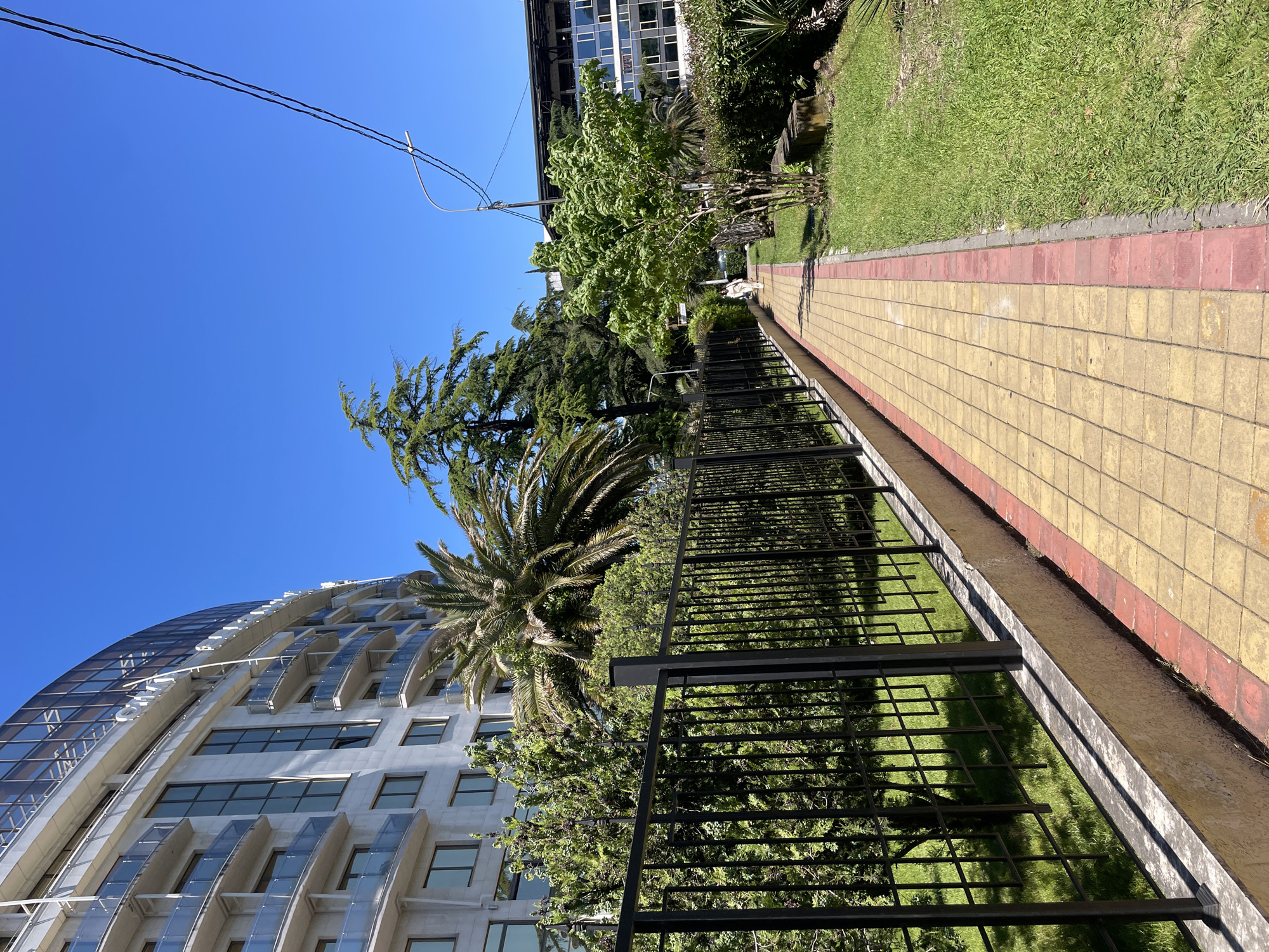 This outdoor scene depicts a paved walkway alongside a green space, with modern buildings under a clear blue sky. The pathway is composed of yellow and red square tiles, extending into the background. A black metal fence separates the walkway from a neatly manicured lawn. Beyond the grass, lush greenery includes a prominent palm tree, tall coniferous trees, and various deciduous trees.
On the left, a striking modern building with a curved facade of glass and light-colored concrete dominates the view, featuring numerous windows and balconies. In the upper right background, another multi-story building with many windows is visible. Utility wires are present against the bright blue sky.
The scene is illuminated by strong sunlight, casting shadows, indicating a clear, sunny day, likely morning or early afternoon. A small, light-colored figure is faintly discernible on the path in the distance. The setting suggests a well-maintained urban or resort area, consistent with the provided context of Sochi, Russia. There is no visible text in the image.