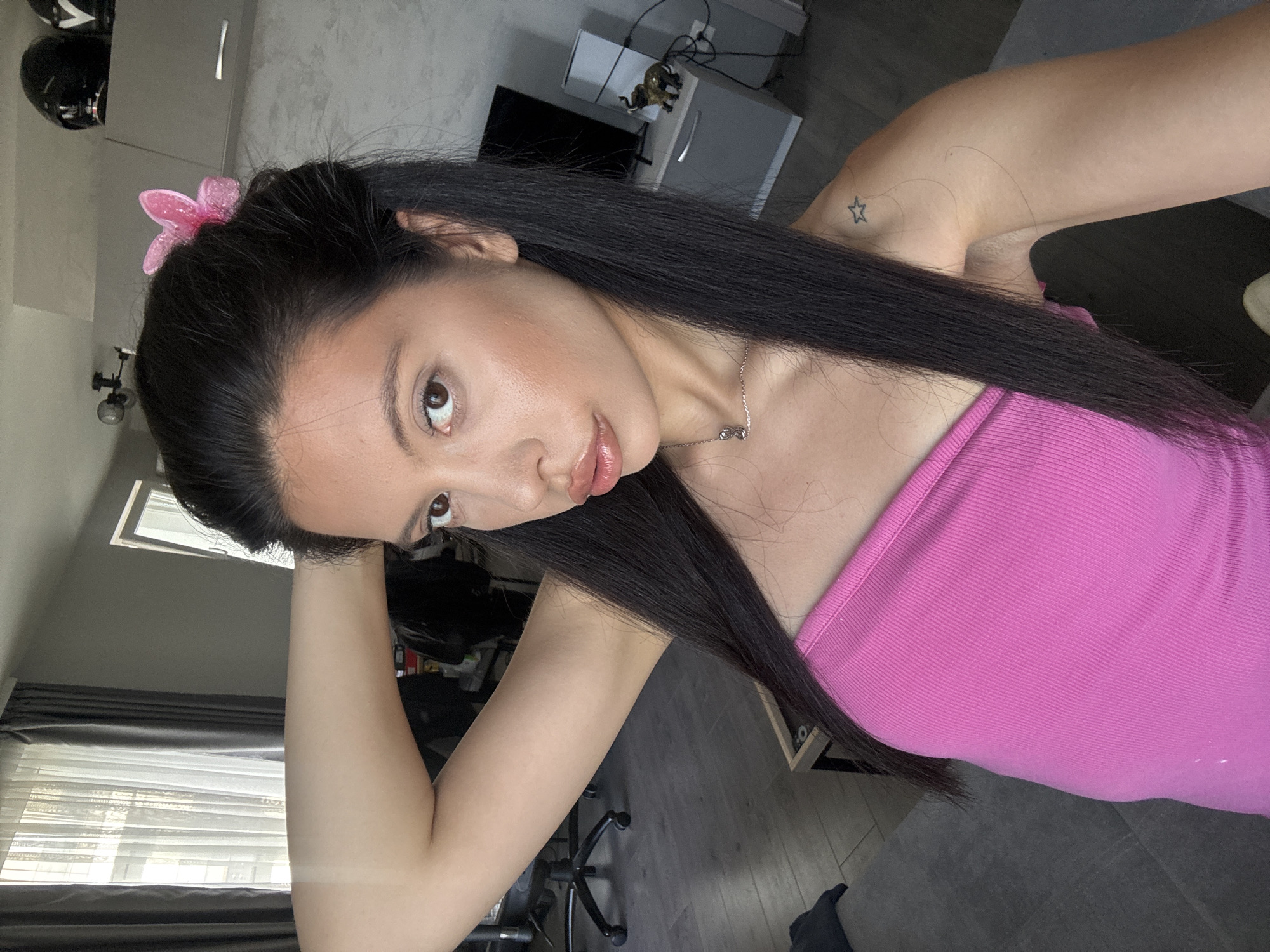 A woman is taking a selfie indoors in what appears to be an apartment. She has long, dark hair with a pink butterfly clip on the side. She is wearing a bright pink strapless top. Her left arm is raised and bent behind her head. A small star tattoo is visible on her right shoulder. In the background, there is a window with curtains, a desk with a chair, a television on a cabinet, and two motorcycle helmets on a shelf. The flooring appears to be wood-like. The lighting suggests daytime. The location is the city of Sofia, Bulgaria.