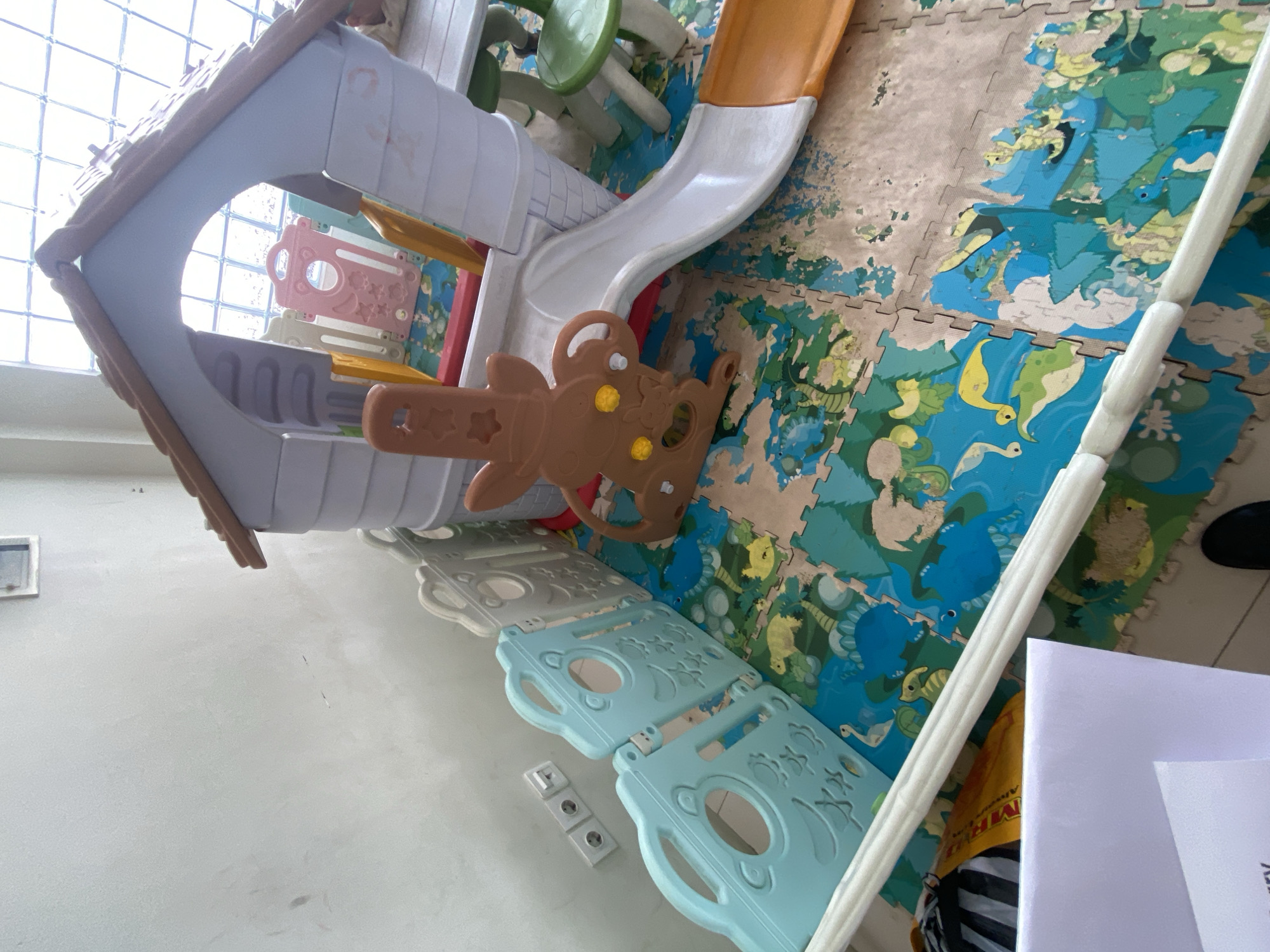The image shows a children's indoor play structure with a slide, a climbing area, and a playhouse. The structure is made of plastic in various colors, including brown, white, gray, pink, and yellow. A brown bear-shaped steering wheel with yellow accents is attached to the side of the play structure. The play area is set up on a mat with interlocking puzzle pieces that have a colorful design featuring cartoon animals like dinosaurs and birds, and a sandy-colored background. In the background, there is a window with a grid pattern, suggesting it might be a glass block window. A white, vertical railing or support is visible on the right side of the image. A white piece of paper with some text is also present in the lower right corner. Electrical outlets are visible on the wall to the left of the play structure. The scene appears to be indoors, likely a playroom or a child's bedroom in Denpasar, Indonesia. The lighting suggests it is daytime, possibly with natural light coming from the window. No people are actively playing in the image, but the presence of the play equipment indicates it is a space for children's activities.
