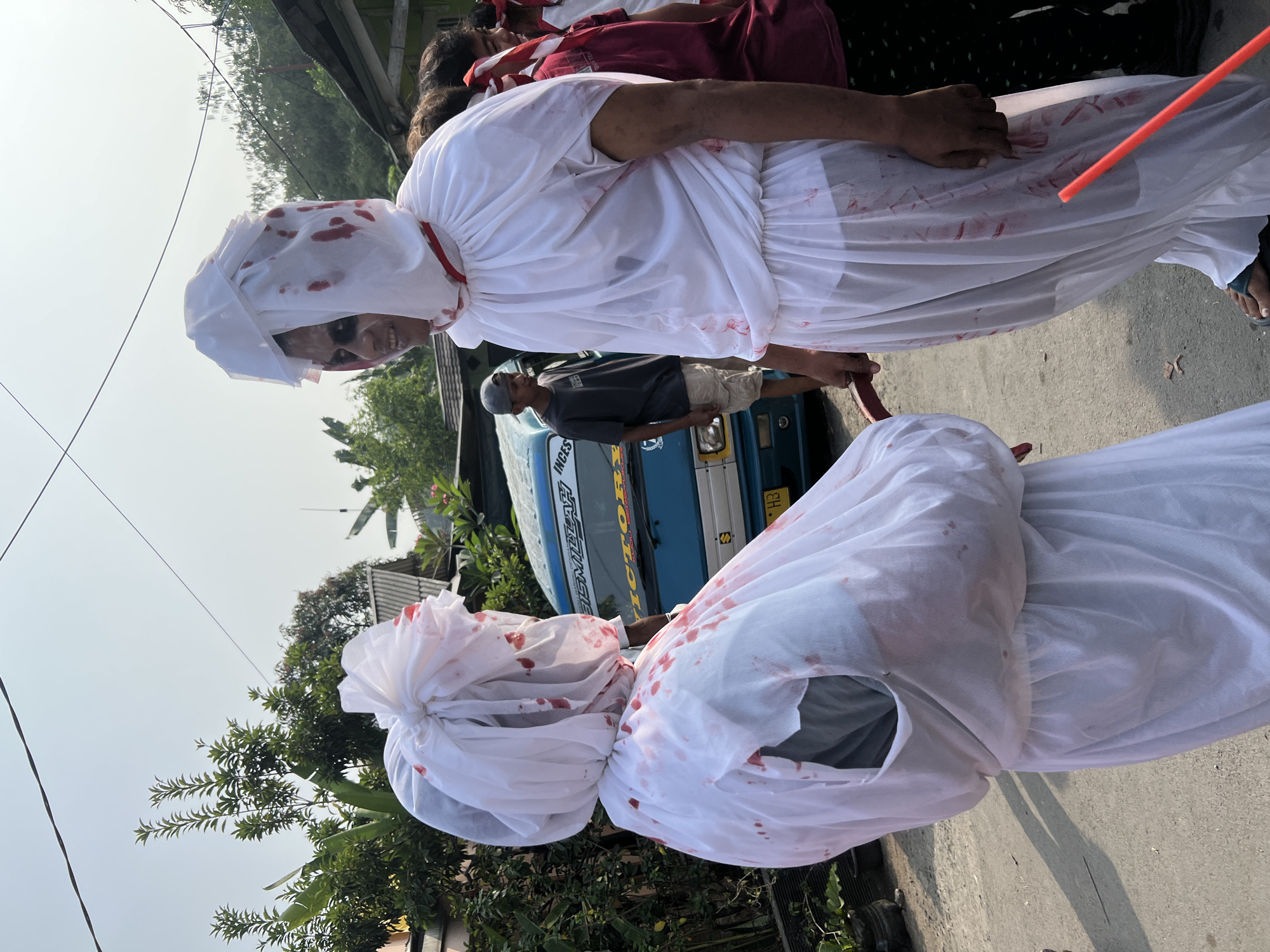 Two individuals, dressed in white costumes resembling ghosts or spirits adorned with simulated blood splatters, stand in a street in Klapanunggal, Indonesia. The costumes consist of flowing white fabric that covers their heads and bodies, with red markings simulating blood. The person on the right has their face visible, featuring dark eye makeup and a grim expression. They are holding a prop resembling a red, curved blade. The person on the left is largely obscured by their costume, with a hole revealing a grey garment underneath. Behind them, a blue truck with text visible on its side is parked. Other individuals, including a person wearing a grey cap and shorts, and others in dark clothing, are present in the background, suggesting a gathering or event. The environment appears to be a residential area with houses and trees visible, under a bright sky.