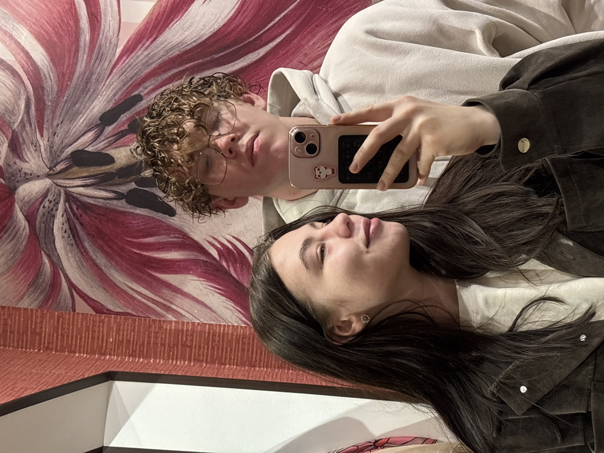 This is a mirror selfie featuring two individuals, a male and a female, in an indoor setting, rotated approximately 90 degrees clockwise.
The male, positioned towards the top of the frame, has curly, light brown hair and wears round, clear-rimmed glasses. He is dressed in a light-colored hooded sweatshirt. He holds a rose gold-colored smartphone, likely an iPhone, with a small white bear sticker on its back case, pointing it towards the mirror for the reflection. His facial expression is neutral.
The female is positioned below the male, with her face and upper torso visible. She has long, dark hair and is wearing a dark brown or grey jacket over a light-colored top. A small, decorative circular earring is visible in her right ear. She has a soft, slightly upward gaze with a subtle, contented expression.
The background is dominated by a vibrant floral pattern on what appears to be wallpaper or a large piece of art. The pattern consists of large, stylized petals in shades of red, pink, and white. Below this floral design, a textured reddish-brown wall or panel is visible. The lower left corner shows a reflection of a white architectural corner.
The activity taking place is a casual mirror selfie. The scene is well-lit, typical of an indoor environment. There are no specific visible cues within the image itself to confirm the provided location context of Perm, Russia. No discernible text is visible in the image.