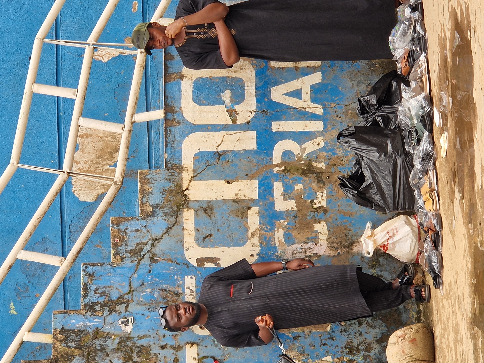 Two men stand in front of a weathered blue wall with white lettering that reads "TECHNICOLOR NIGERIA". The man on the left, wearing a black pinstripe tunic and sunglasses perched on his head, holds something orange in his hand and gestures with it. He has a watch on his left wrist and wears black sandals. The man on the right, in a black tunic and a green baseball cap, has his arms crossed and his right hand touching his chin. He wears a bracelet on his left wrist. In front of them, on the ground, are several pairs of shoes and sandals, some in plastic packaging, along with two large black plastic bags and a white plastic bag. A large, light-colored rounded object, possibly a large stone or bag of material, sits to the left of the men. The ground appears wet in places, reflecting the surrounding environment. The scene is outdoors, likely on a street or market.