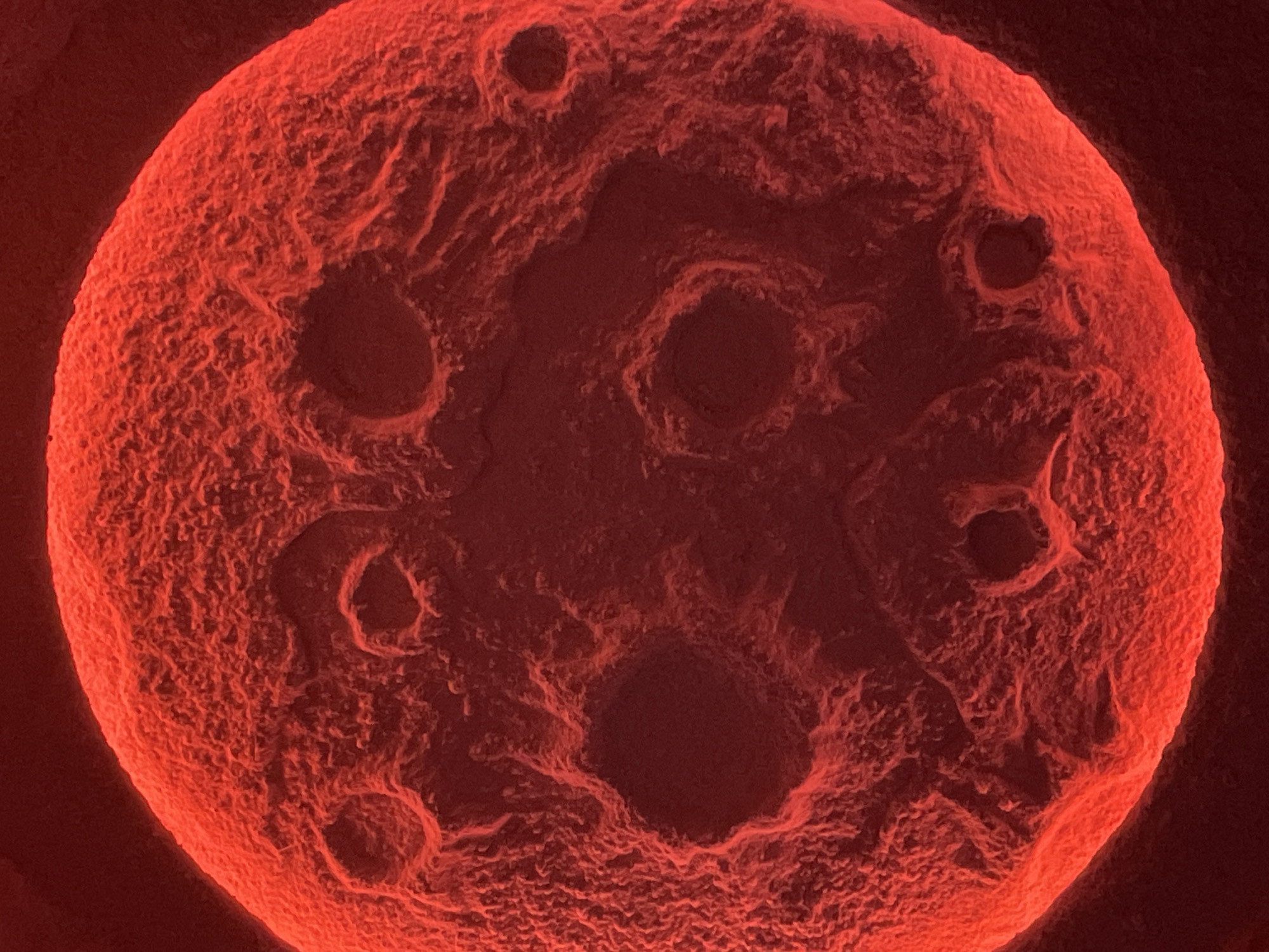 The image displays a highly magnified, irregularly spherical object, predominantly rendered in shades of red and dark red against an unidentifiable black background. The object's surface is heavily textured, appearing rough, porous, and undulating, with numerous depressions, pits, and crater-like formations of varying sizes. Brighter red highlights illuminate the raised contours and edges, particularly on the upper and left sides of the sphere, suggesting a directional light source. Deeper recesses and shadowed areas are depicted in darker crimson tones, approaching black. The object occupies the majority of the frame, presented in extreme close-up. No people, other distinct subjects, objects beyond the primary sphere, actions, or interactions are visible. The surrounding environment is an undifferentiated void. The image provides no visual information to determine its geographic location, including within the city of Vinnytsia, Ukraine.