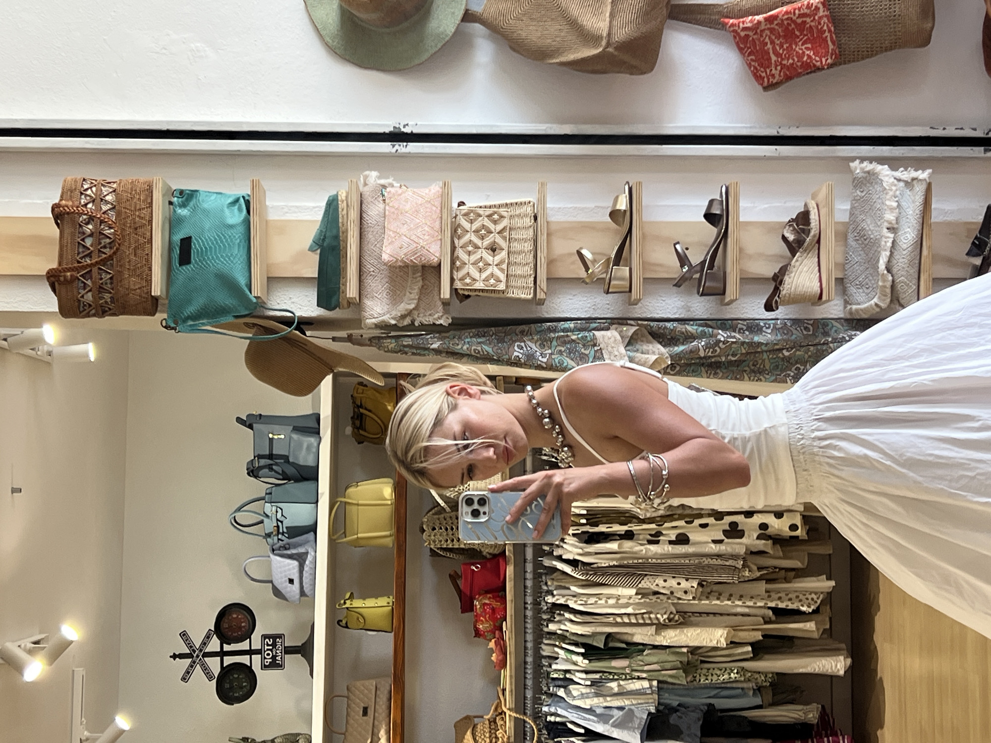 A young blonde woman, wearing a white tank top and a white full skirt, stands in a boutique and takes a mirror selfie with her phone. She is accessorized with a chunky silver necklace and several silver bracelets on her left arm. In the background, the boutique displays clothing on racks and accessories such as handbags, hats, and shoes on shelves. The scene is well-lit with overhead spotlights. The location is a boutique in Pollença, Spain.