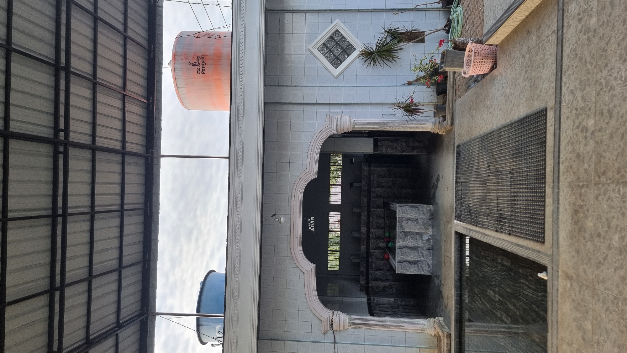 The image captures an outdoor area of a building, likely a public facility in Meunasahlhok, Indonesia. In the foreground, a dark pool of water is visible to the left, bordered by concrete. To the right of the pool is a dark grey textured mat. Behind these, an arched entryway leads into a dimly lit interior where rows of water faucets are visible, suggesting a bathing or ablution area. The entryway is flanked by decorative pillars. The exterior walls of the building are tiled in a light blue, with a diamond-shaped decorative element on the right wall. Above the entryway, text reads "KAUM ADAM", likely indicating a men's section. On the right side of the building's exterior, there are potted plants. In the background, two large water tanks are visible on the roof, one blue and one orange with the "Penguin" logo. The sky is overcast.