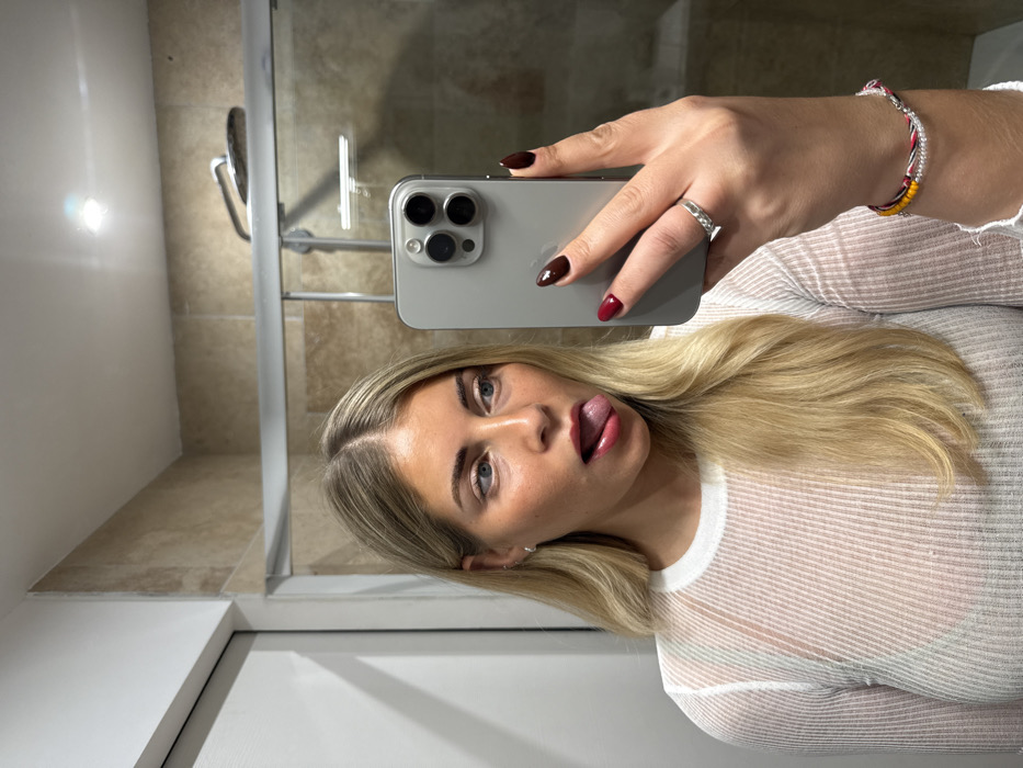 A blonde woman takes a selfie in a bathroom. She holds a grey smartphone with manicured hands; one hand wears a silver ring and a colorful bracelet. The woman is sticking her tongue out with a playful expression. She's wearing a sheer, white, long-sleeved top over what seems to be a dark undergarment. A shower with a glass door and tiled walls is behind her. Part of a white door frame is also visible in the lower left corner.