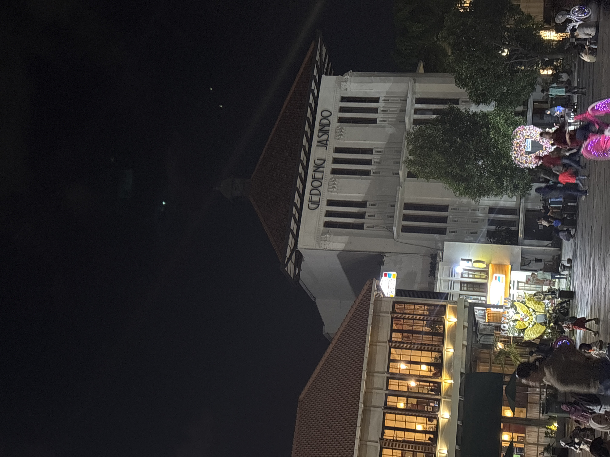 The image depicts a night scene in Fatahillah Square, a public square in the Old Town of Jakarta (Kota Tua), West Jakarta, Indonesia. In the background, a large white building with the prominent inscription "GEDOENG JASINDO" is illuminated. Adjacent to it, to the left, is another building with a reddish-brown tiled roof and windows with warm interior lighting. The square itself is paved and populated by a moderate number of people. Several individuals are visible walking, standing, and sitting. In the mid-ground, there is a large, ornate floral decoration, possibly a wreath or display, with a heart shape within it. To the right of the floral display, a person is riding a brightly lit pink bicycle. Various smaller stalls or shops with illuminated signs are also present, including one with the "SPARK" logo. The sky above is dark, with faint celestial lights visible. The overall atmosphere suggests an evening gathering or event in a historical urban setting.