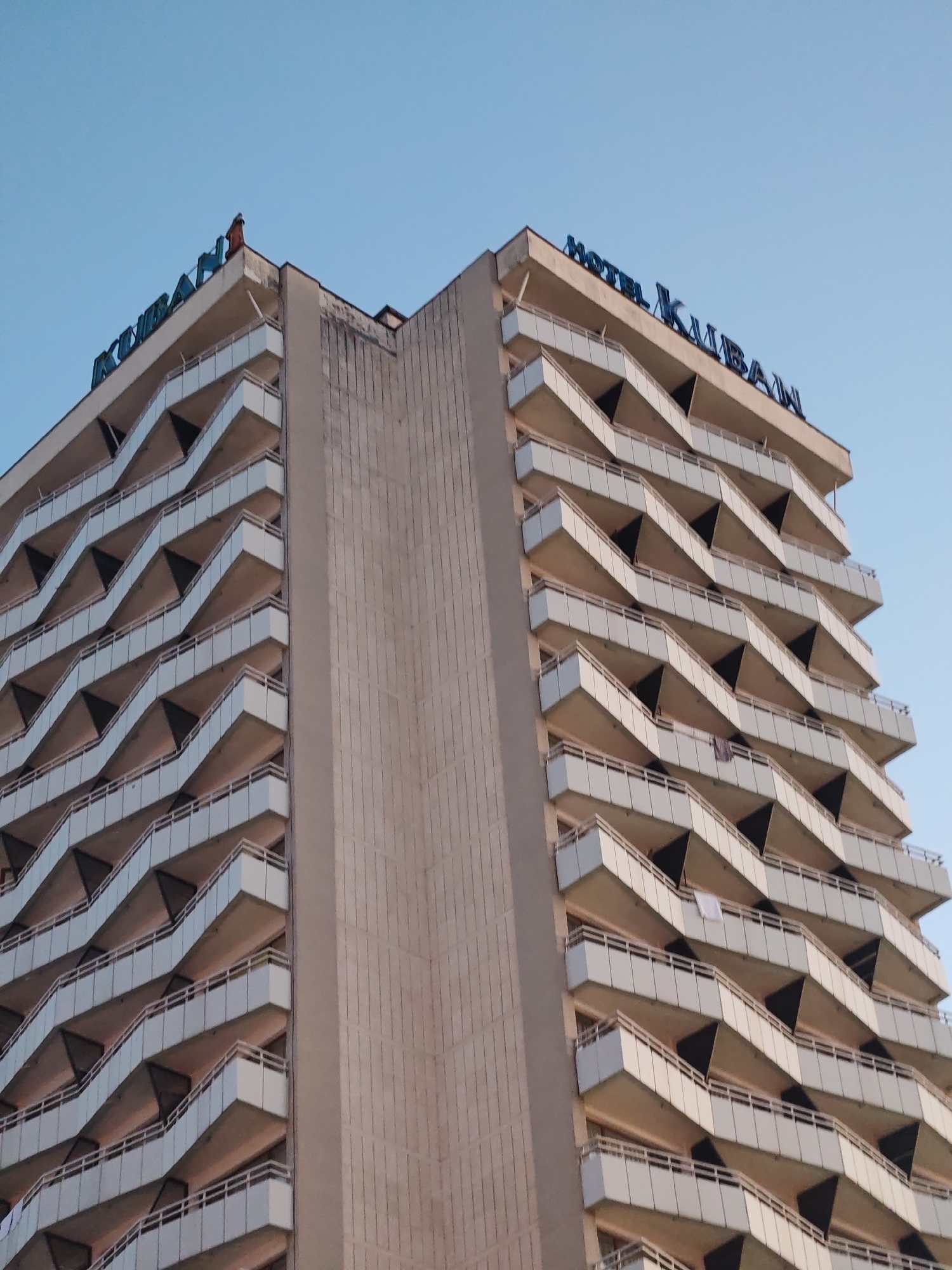 The image shows a tall building with a unique architectural design. It is a hotel called "Hotel Kuban," as indicated by the large blue lettering on the top. The building has numerous floors, each with a protruding balcony that creates a staggered, zigzag pattern. The central part of the building is a flat, unadorned concrete slab. The sky is a clear blue. A small figure appears to be standing on the roof of the hotel. A white cloth is hanging on one of the balconies. The building is located in Nesebar, Bulgaria.