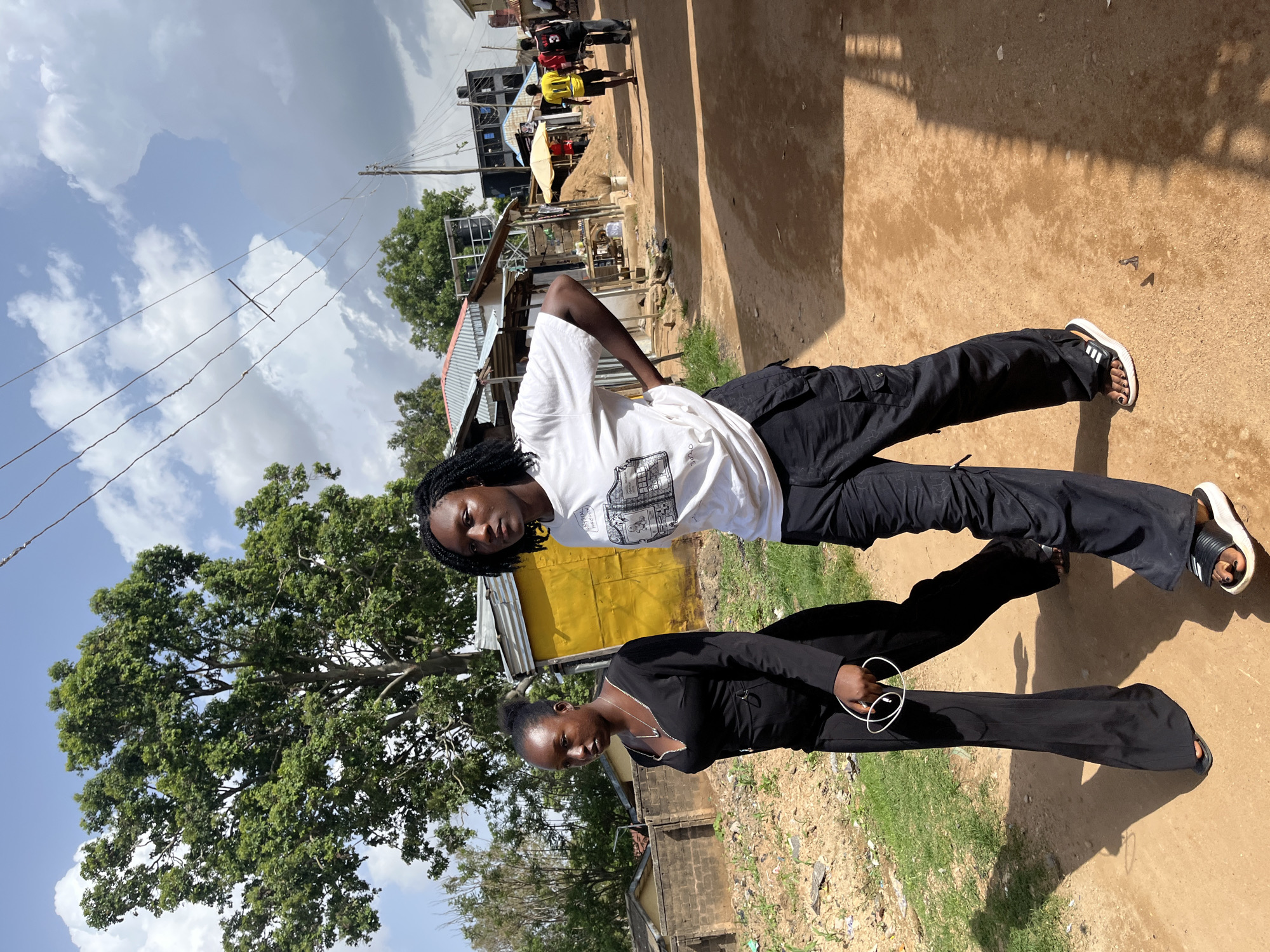 In Rafin Par, Nigeria, two young women walk on a dirt path beside a row of buildings. The woman on the left wears a black, long-sleeved dress with a V-neckline and holds a white cord in her hand. She has dark skin and her hair is pulled back into a bun. The woman on the right has dark skin and her hair is styled in braids. She wears a white t-shirt with a graphic print and black cargo pants. They walk towards the viewer, with the woman on the right looking directly at the camera. In the background, there are buildings with corrugated metal roofs and walls, some painted yellow. A large tree with green foliage is visible to the left of the frame. Other people and structures are visible further down the path. The sky is partly cloudy.
