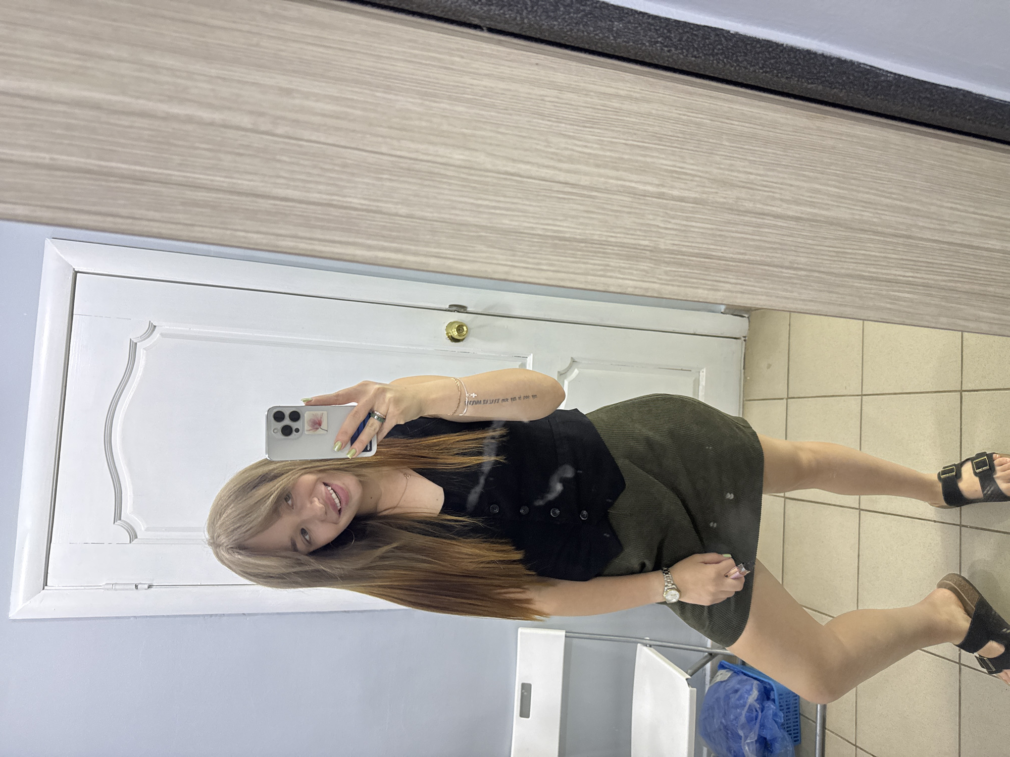 A young woman with long, blonde-brown hair and bangs is taking a mirror selfie indoors. She is smiling and looking at her phone, which is held up to take the picture. She is wearing a black vest over a black v-neck top and a dark green corduroy skirt. On her left wrist, she wears a silver watch. She has a tattoo on her right forearm and is wearing rings on her fingers. She is also wearing black Birkenstock sandals.
The background shows a white door with decorative trim and a gold doorknob, a pale blue wall, and a portion of a wooden closet or cabinet. To the left of the woman, there is a white folding chair and a blue plastic bin with a blue bag inside. The floor is tiled with light beige square tiles. The image was taken in an indoor setting, likely a dressing room or a similar space, within the city of Chaikovsky, Russia.
