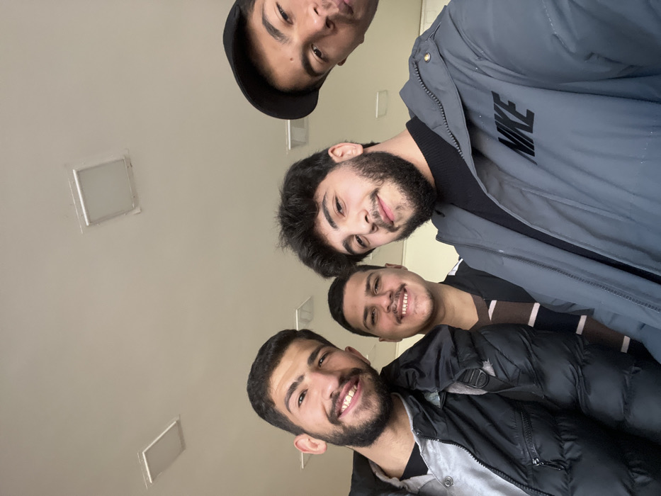This image captures a selfie of four young men indoors in the city of Rukn ad-Din Municipality, Syria. The scene is illuminated by artificial light from two rectangular flush-mounted ceiling fixtures on a light-colored ceiling, which occupies the upper half of the frame.
From left to right:
1. A young male with dark hair and a short beard is smiling. He wears a black puffer jacket over a light grey collared shirt. A black strap is visible on his left shoulder.
2. Slightly behind the first individual, another young male with short dark hair and a mustache is smiling. He is wearing a dark, possibly striped, top.
3. In the foreground, a young male with dark wavy hair and a full beard is smiling. He wears a grey puffer jacket with the word "NIKE" printed in black on the right chest area, over a black ribbed shirt.
4. Partially visible on the far right, a fourth young male is present, with only the upper portion of his face and a black baseball cap visible. He is looking directly at the camera.
All individuals appear to be in their late teens or early twenties and are positioned closely together, facing the camera.