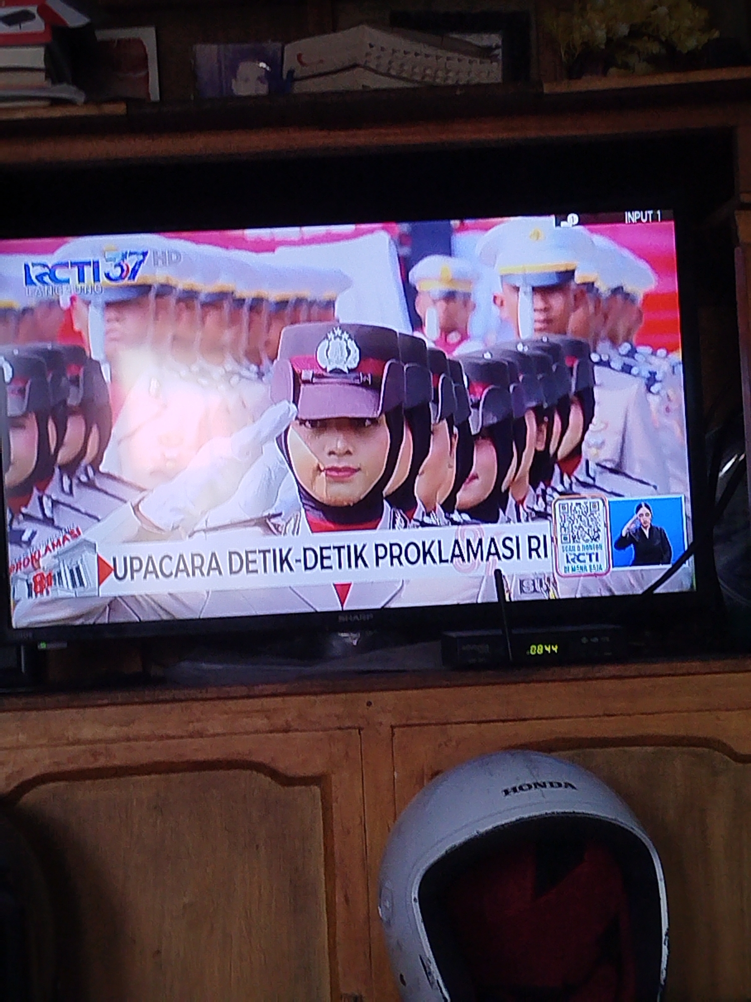 The image shows a television screen displaying a news broadcast. The broadcast features a woman in a police uniform, saluting. The text on the screen reads "UPACARA DETIK-DETIK PROKLAMASI RI". This translates to "Ceremony of the Moment of Proclamation of the Republic of Indonesia," referring to Indonesia's Independence Day. The broadcast is from RCTI 37 HD, an Indonesian television channel. The television is placed on a wooden cabinet, and a white Honda helmet is visible in the foreground. The background suggests an indoor setting with wooden furniture and some decorative items on a shelf above the television. There is no visible indication of the city of Balikpapan, Indonesia, in the image.