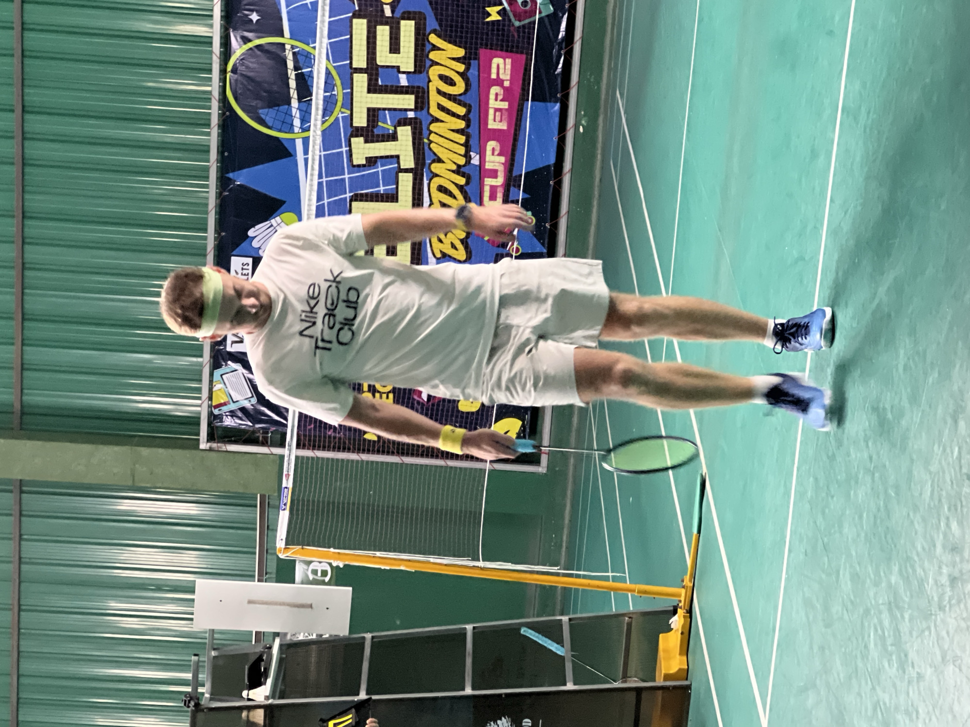 A young man is playing badminton indoors. He is wearing a white t-shirt with "Nike Track Club" printed on it, light gray shorts, a light green headband, and blue badminton shoes. He is holding a blue badminton racket and is in the middle of a game. The court is green with white lines. In the background, there is a banner that reads "ELITE BADMINTON CUP EP.2". The walls of the facility are green. The image appears to be taken at a badminton court in Ban Rang Rak, Thailand.