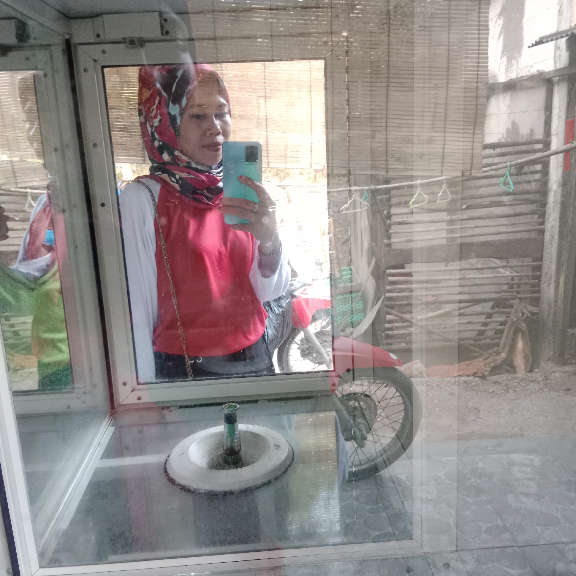 A woman in a red and white shirt and a colorful hijab takes a selfie in a glass display case. She is holding a teal smartphone in her right hand. The display case is made of metal and glass, and it appears to be in an outdoor market or a roadside stall. In the background, a red motorcycle is parked. Behind the motorcycle, there is a wooden wall covered with a woven mat. The ground is a mixture of dirt and gravel. The overall scene suggests a casual, everyday setting in Tegowanu Kulon, Indonesia.