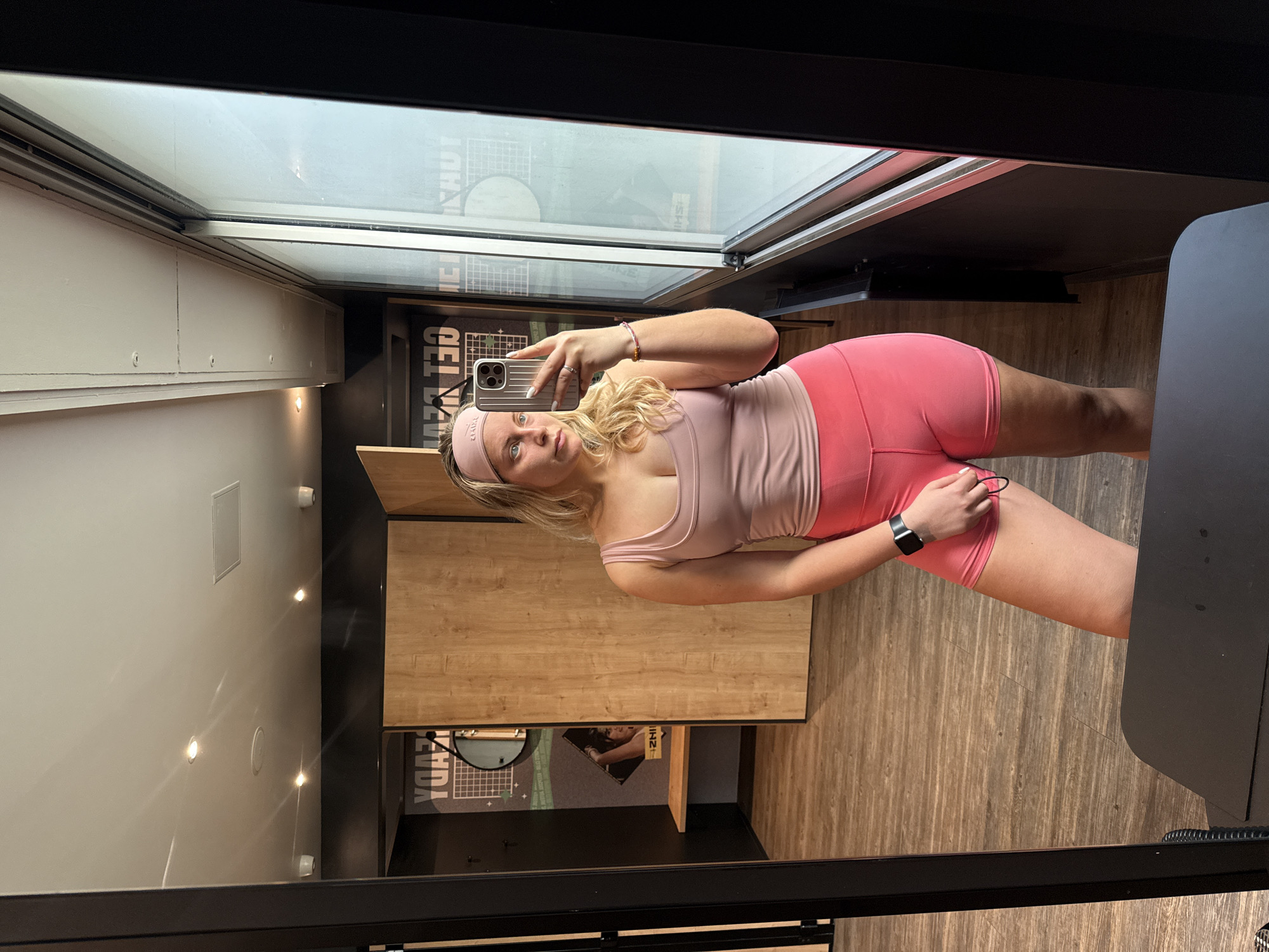 The image depicts a woman taking a selfie in a room that appears to be a locker room or dressing area. She is wearing a light pink tank top and matching shorts, with a pink headband. A black smartwatch is visible on her left wrist. She is holding a cell phone in her right hand, likely to take the picture. The room has wooden panels, mirrored walls, and a window. A piece of furniture sits to the right. A poster is visible on the wall. The presence of the poster and mirror suggests the location is not only a changing area, but a business that wishes to promote itself. However, nothing indicates this is a Parisian location.