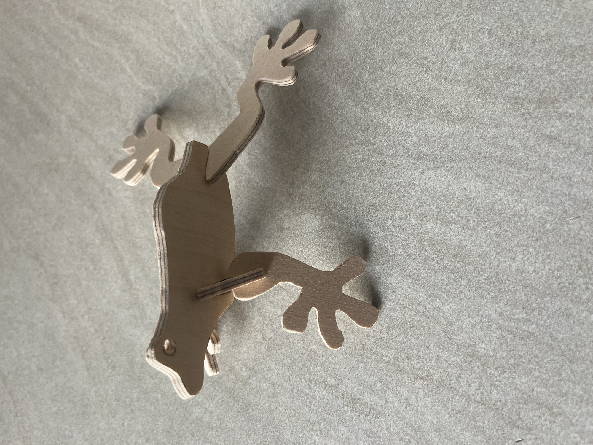 The image shows a light brown, wooden, three-dimensional cut-out of a stylized gecko or lizard. It is positioned on a light grey, speckled surface, likely a countertop or textured wall. The gecko is oriented vertically in the frame, with its head pointing downwards towards the bottom left corner. Its body is flat, and its limbs are articulated and spread out, suggesting it is either climbing or in a resting position. The gecko has four limbs, each ending in five finger-like digits. A small circular hole is visible near the head, possibly for hanging or as part of the design. The lighting casts subtle shadows, indicating the light source is from the upper right. The overall scene is simple and focused on the object, with no other prominent subjects or background details. There is no visible text in the image. The location context is Jelgava, Latvia, but there are no specific visual cues in the image to confirm this.